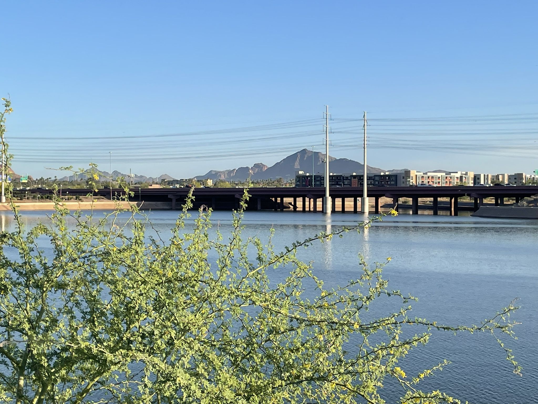 Lake with a bridge, distant mountains, and buildings under a clear sky. - Rio Salado Pathway