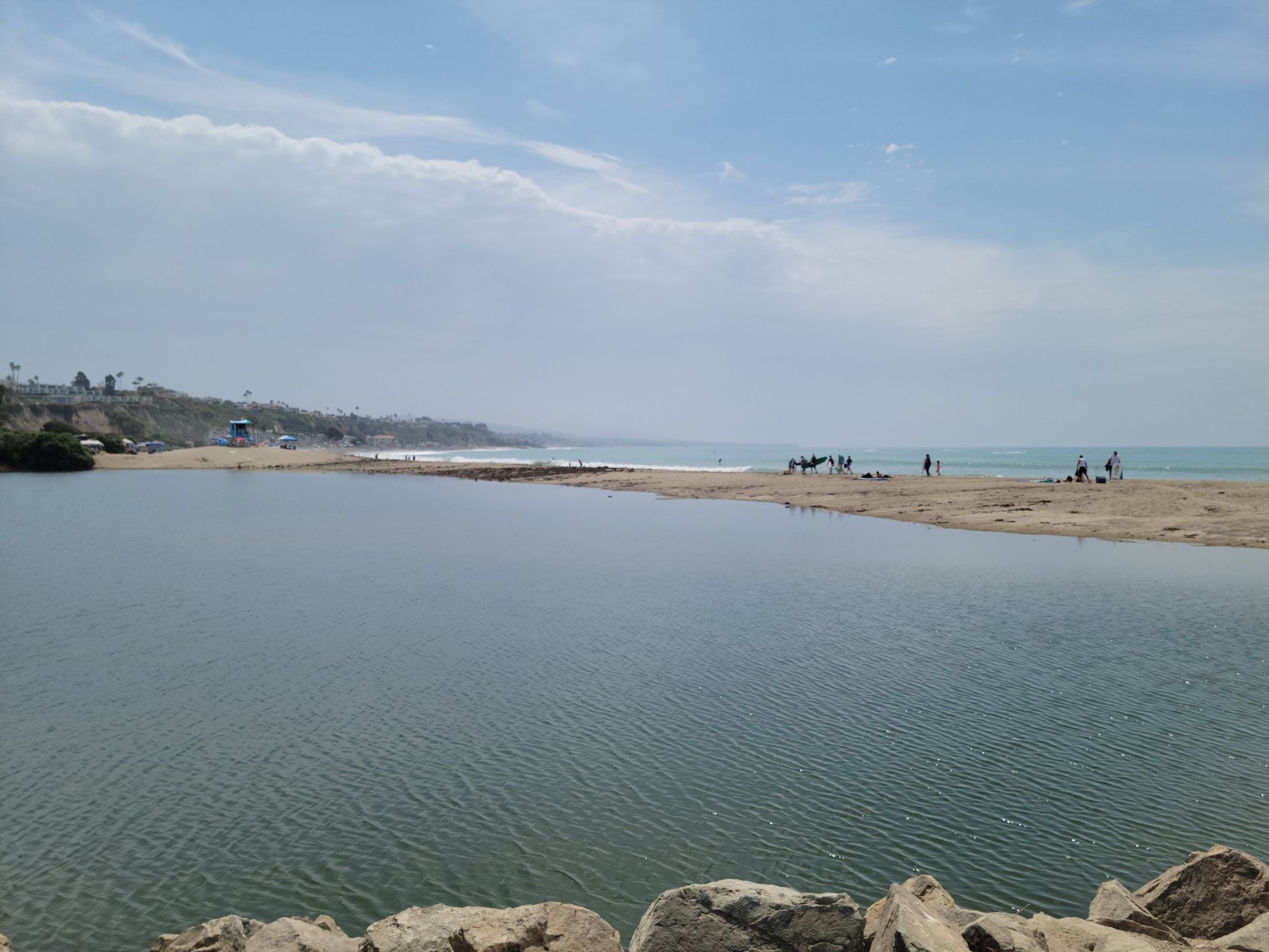Calm lagoon meeting ocean, people on sandy beach under a partly cloudy sky. - San Juan Creek Trail