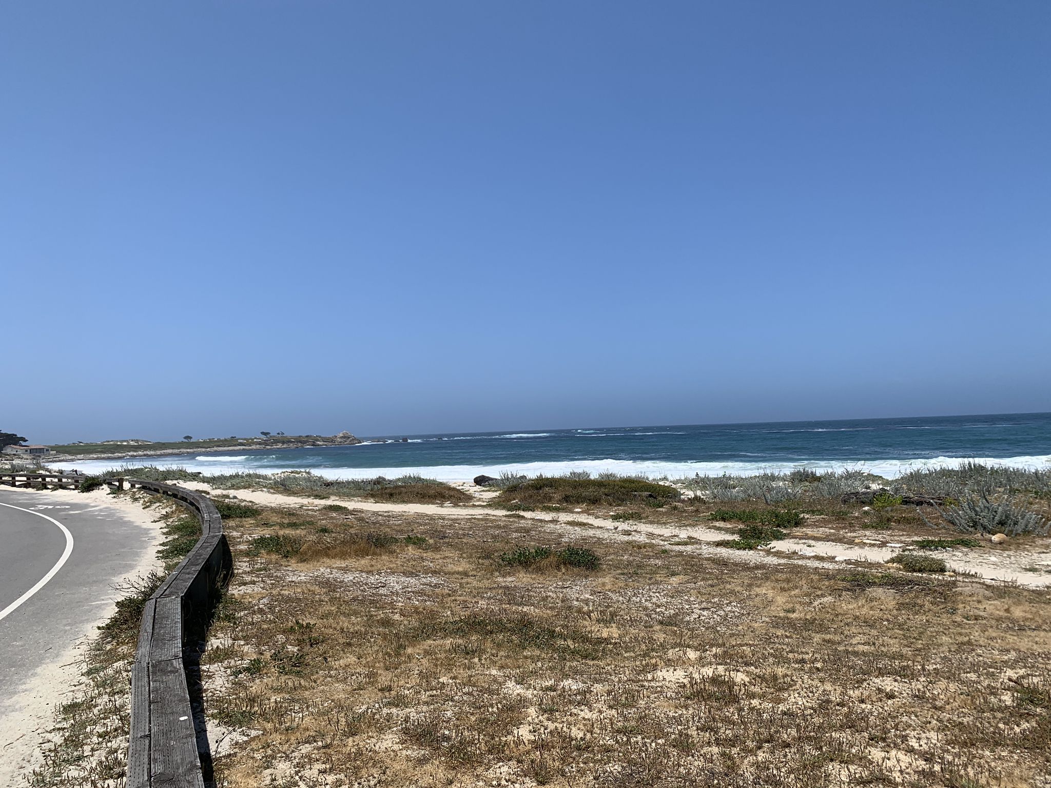Sunny coastal view with crashing waves, a sandy dune, and a winding road. - Spanish Bay Trail