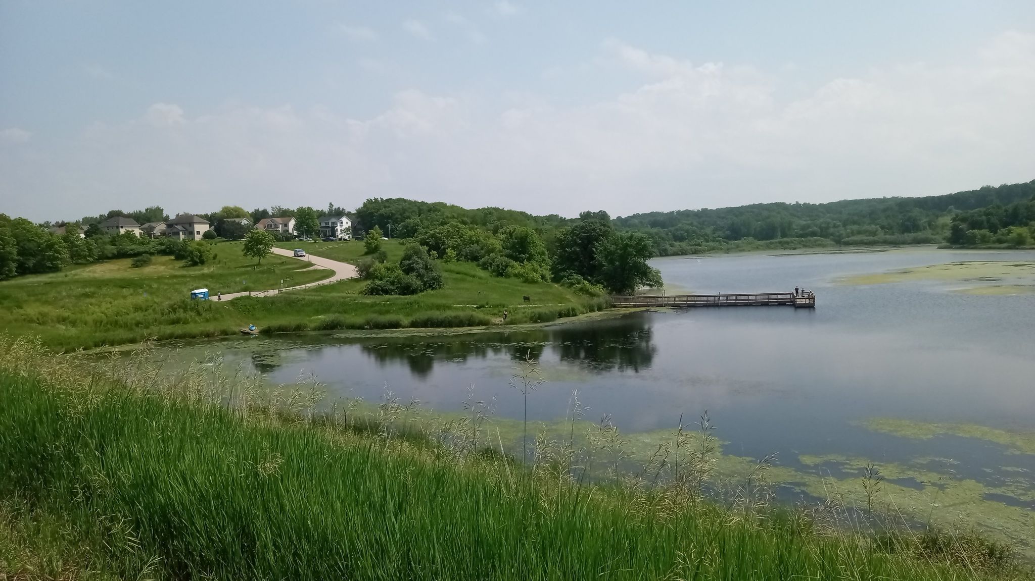 A calm lake with a long dock, surrounded by green hills and distant houses. - Willow Creek Reservoir Loop