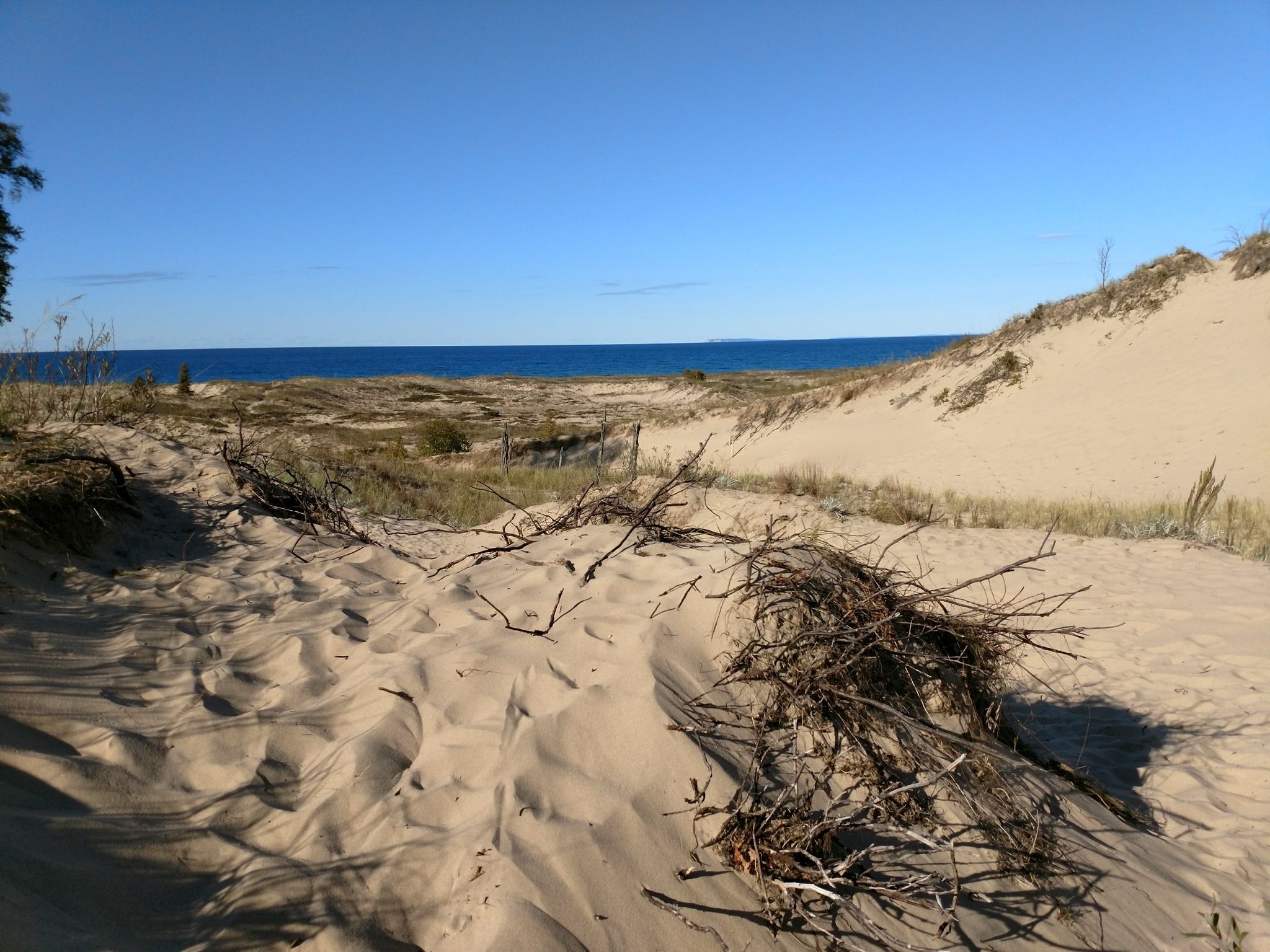 Sandy dunes with dry brush leading to a vast blue body of water. - Old Indian Trail