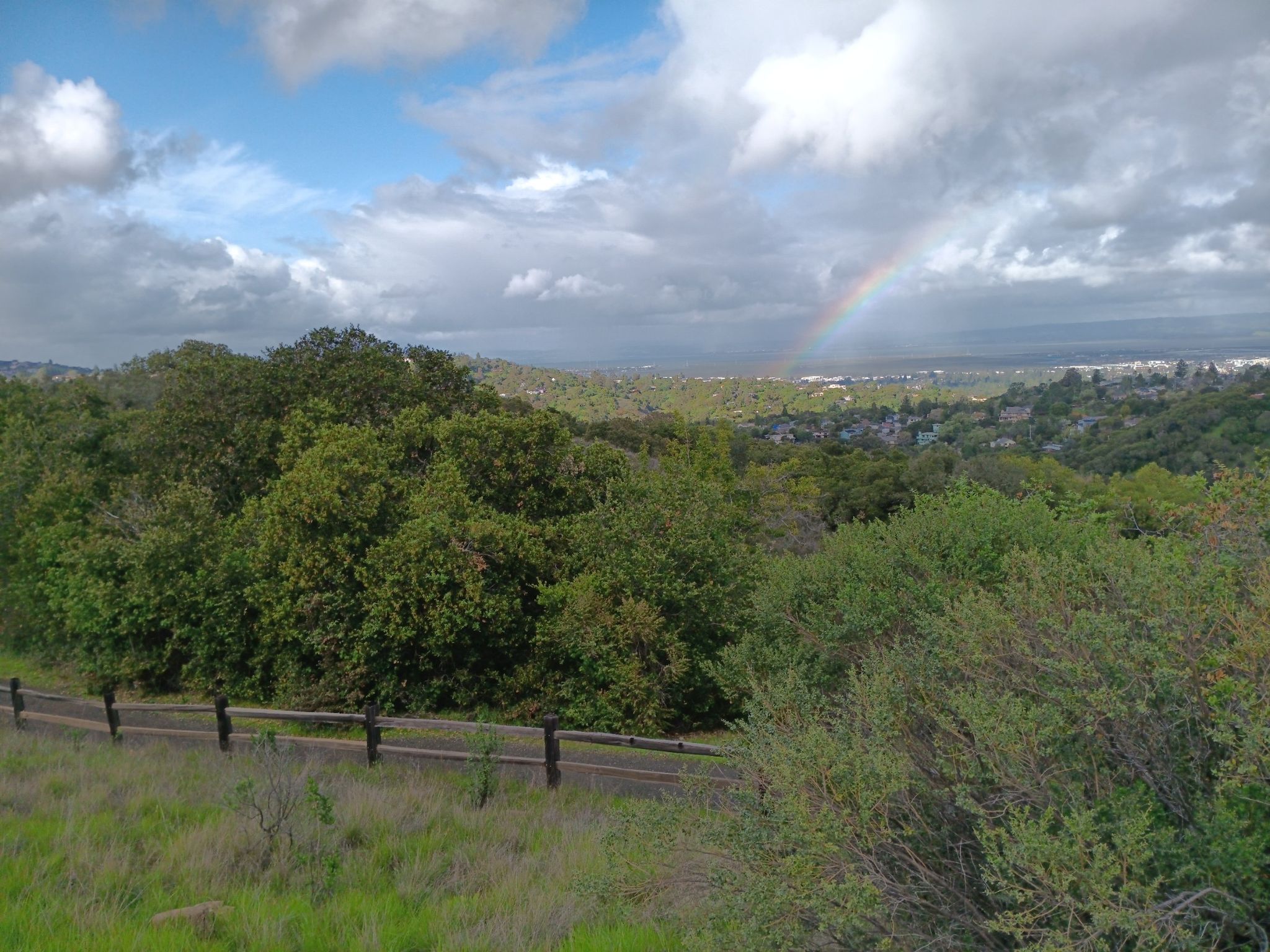 A vibrant rainbow arches over a lush green valley with distant buildings. - Sylvan Loop