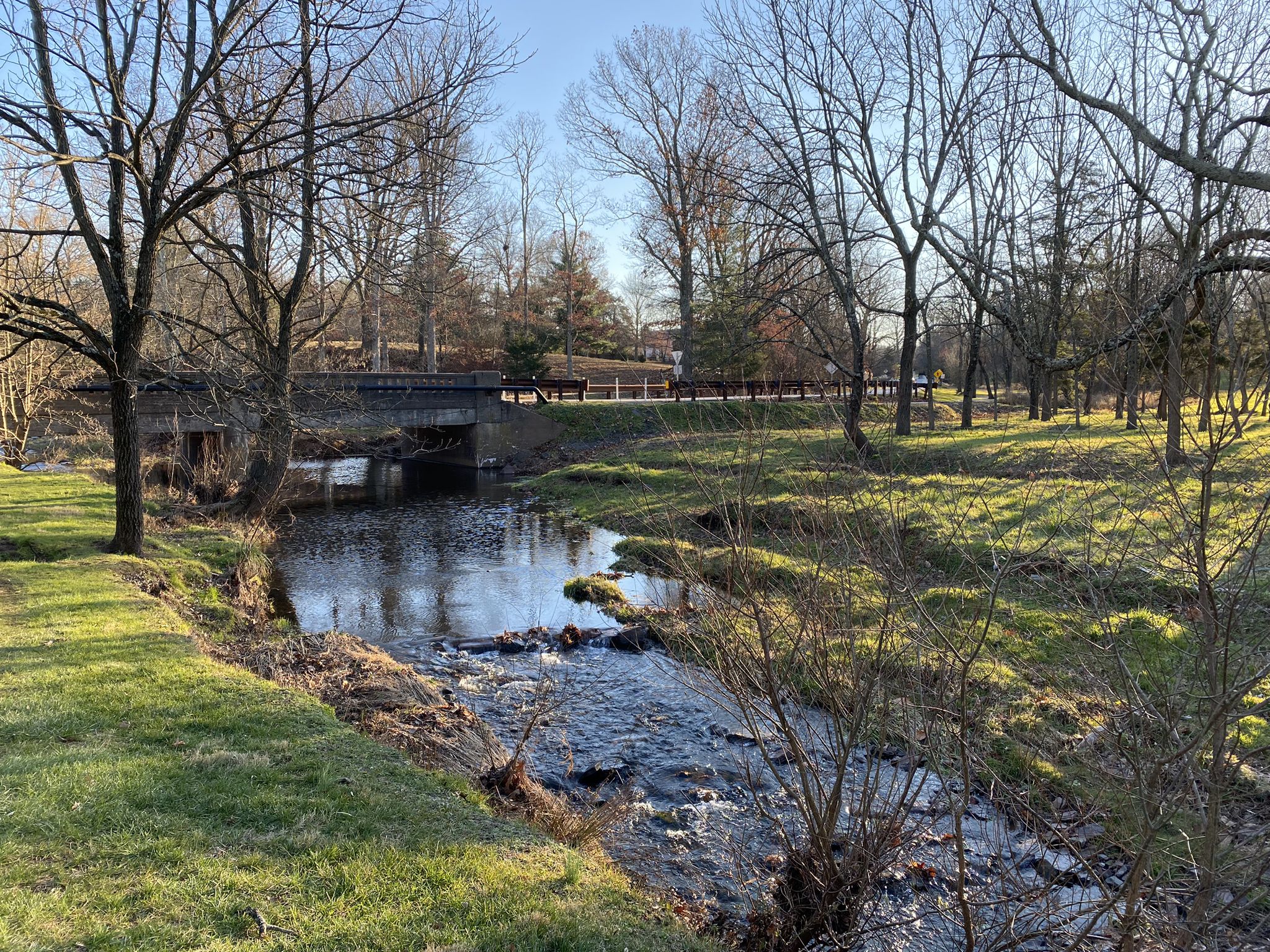 A rocky stream flows under a bridge, surrounded by grassy banks and bare trees. - Zacharias Creek Trail