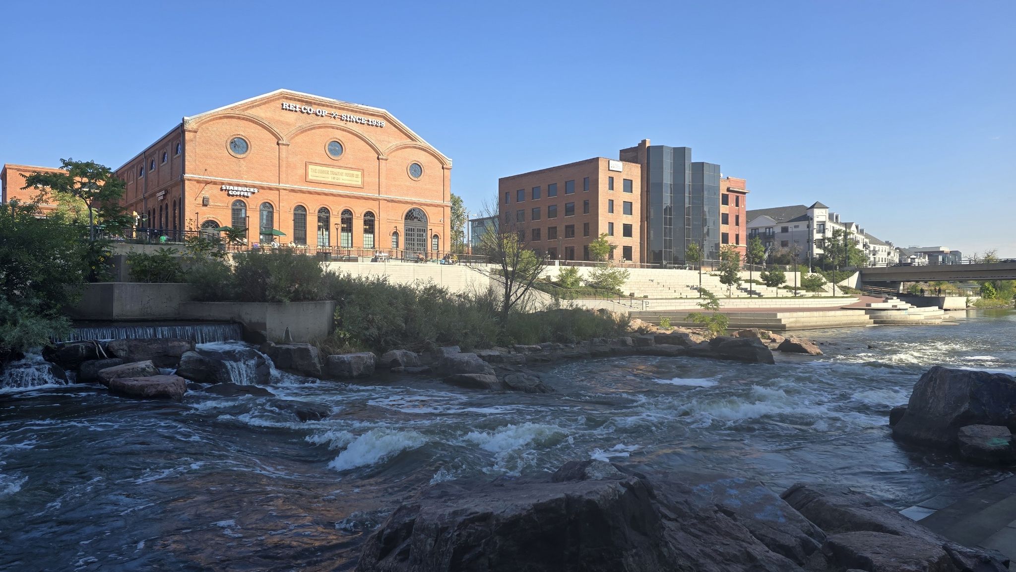 A wide river with rapids flows past a large brick building under a clear sky. - Washington Park Flower Gardens – Cherry Creek Trail loop from Glendale