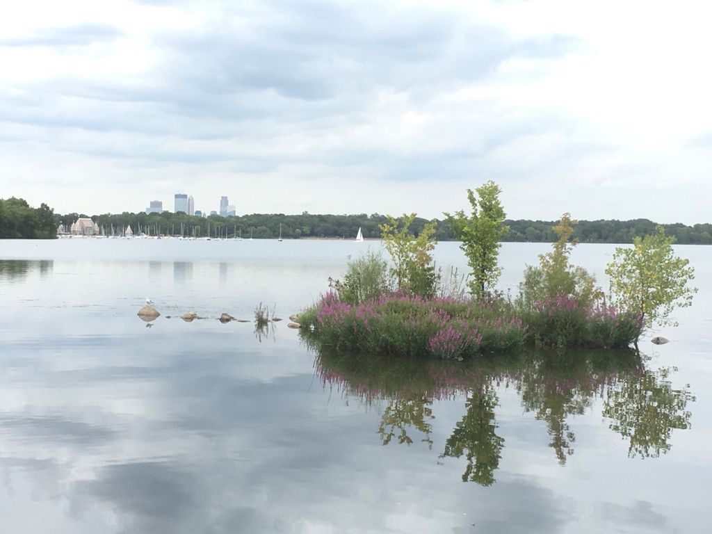 Small island with pink flowers and trees on a lake under a cloudy sky. - Lake Harriet – Bde Maka Ska loop from Southwest