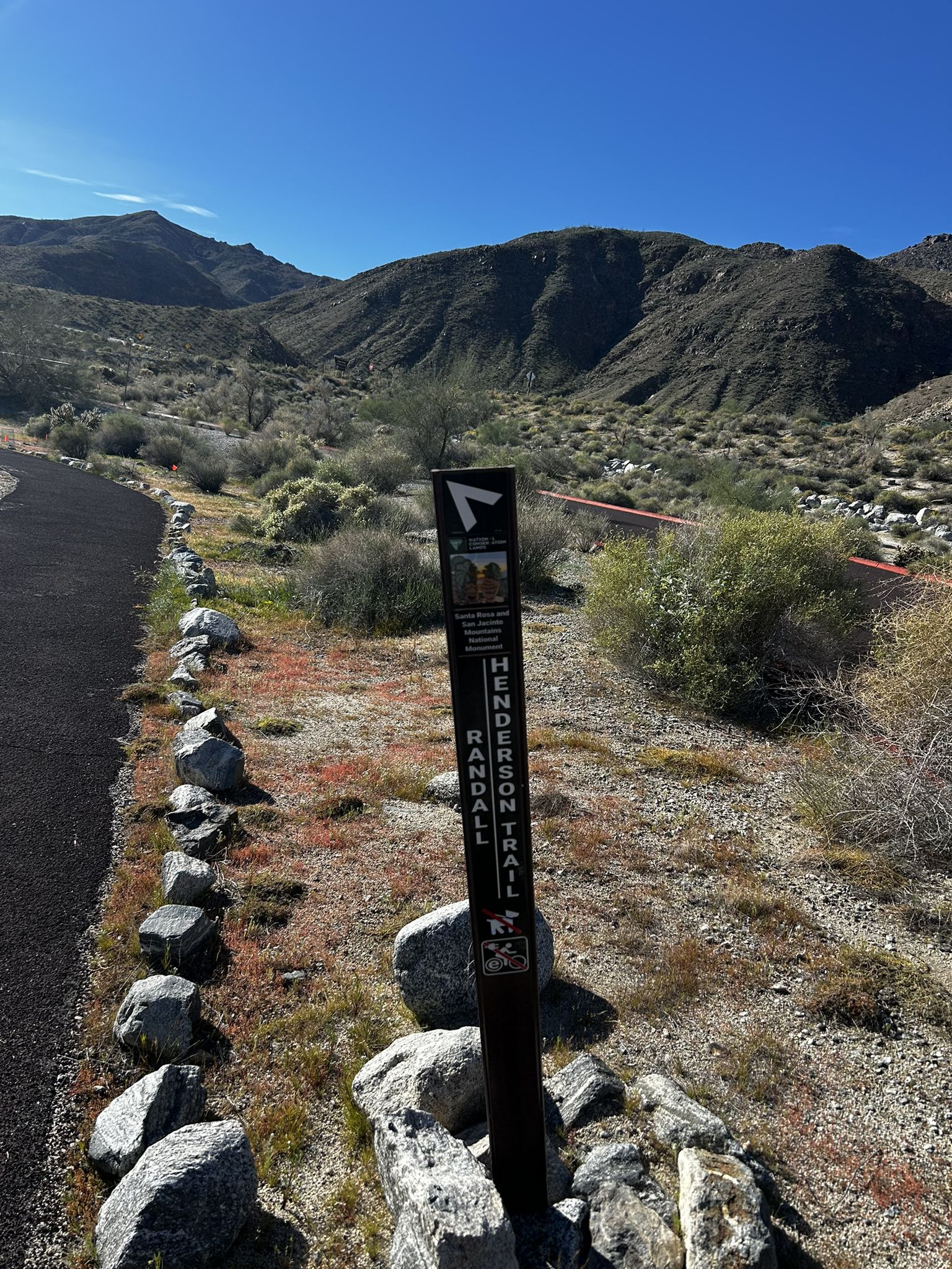 A trail marker sign stands beside a paved path in a desert landscape. - Art Smith Loop