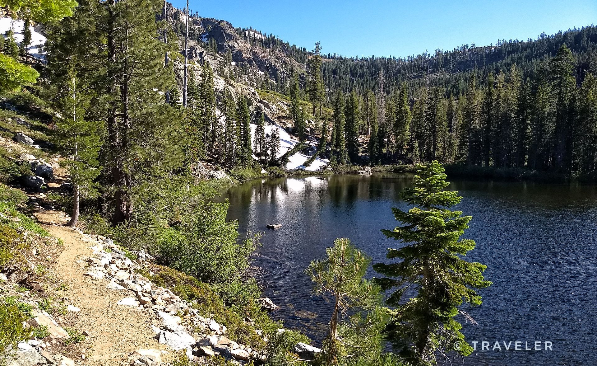 Clear lake with snowy banks, surrounded by pine trees and a dirt path. - Pacific Crest Trail to Tamarack Lakes loop