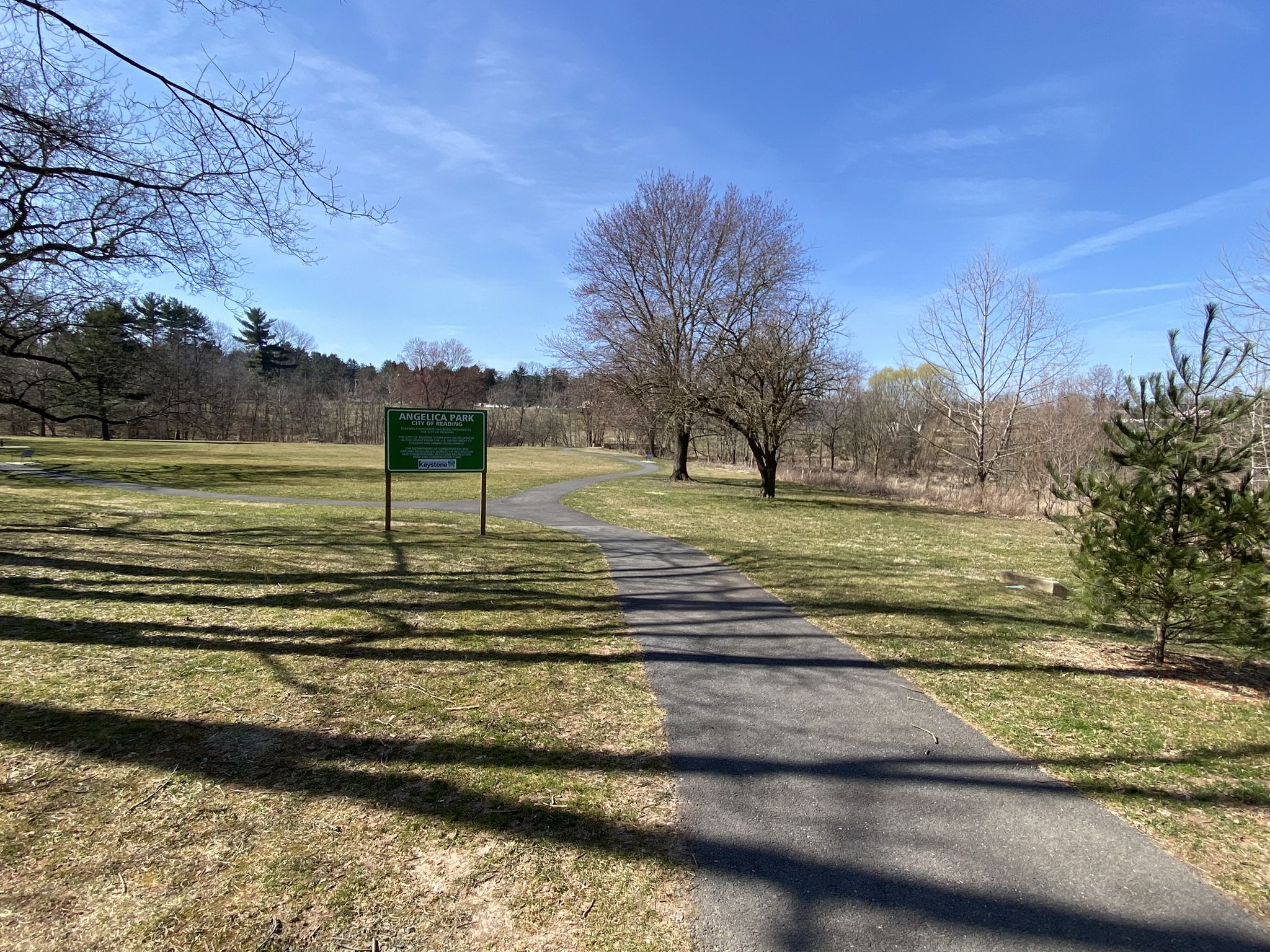 Paved path through a sunny, grassy field with bare trees and a green sign. - Mohnton loop