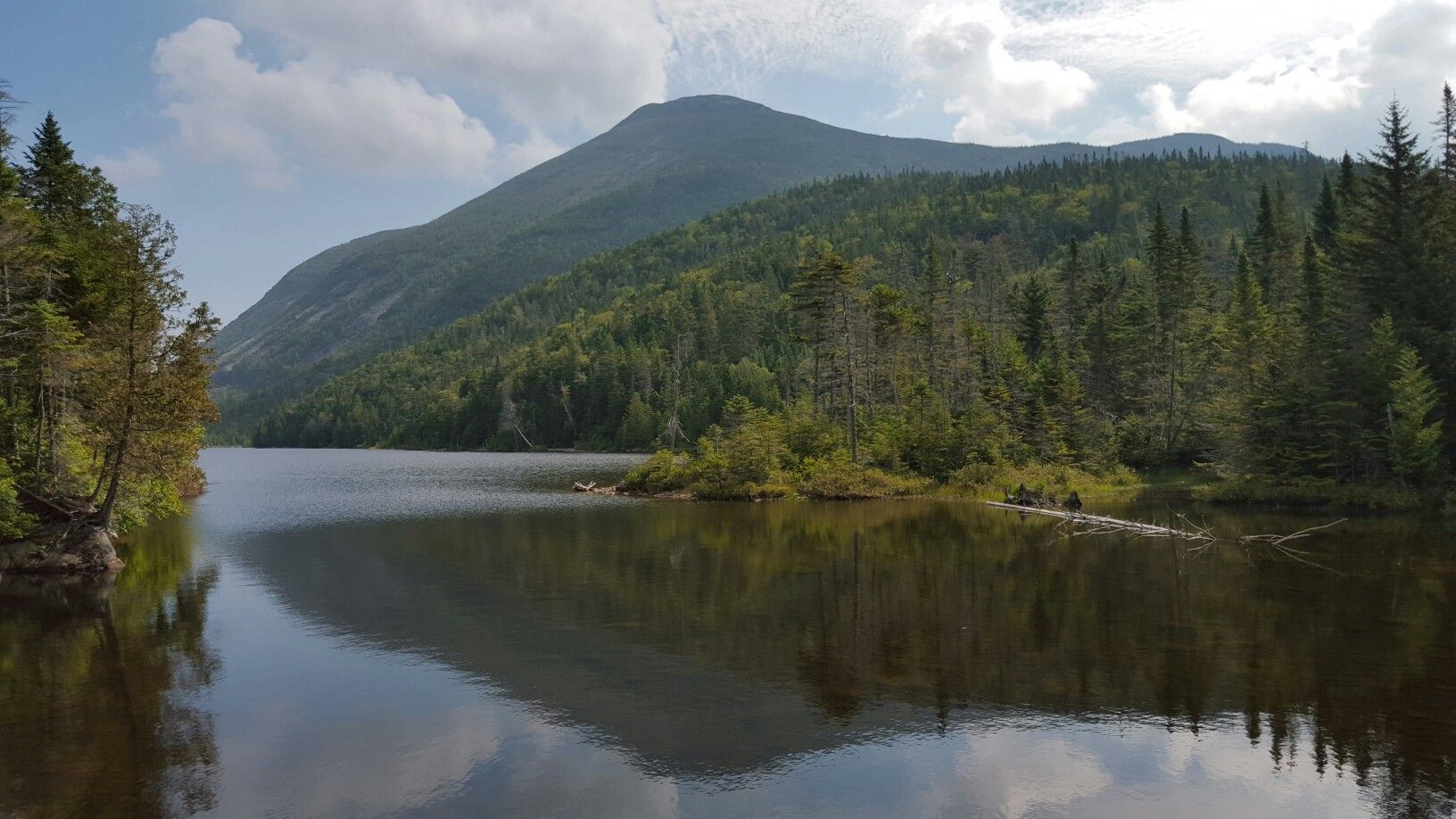 Lake reflecting a large forested mountain under a partly cloudy sky. - Calamity Brook Trail