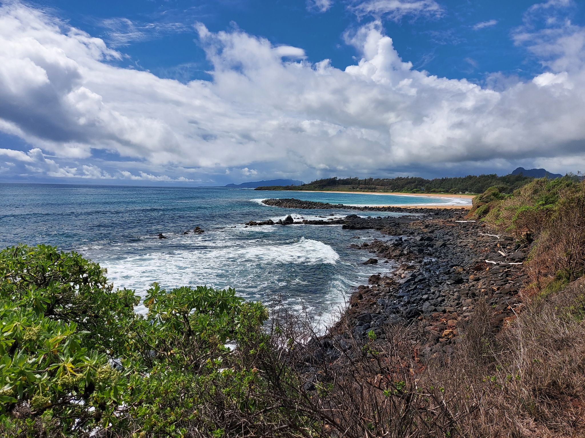 Rocky coastline with crashing waves, distant sandy beach, and cloudy sky. - Ke Ala Hele Makalae Coastal Loop