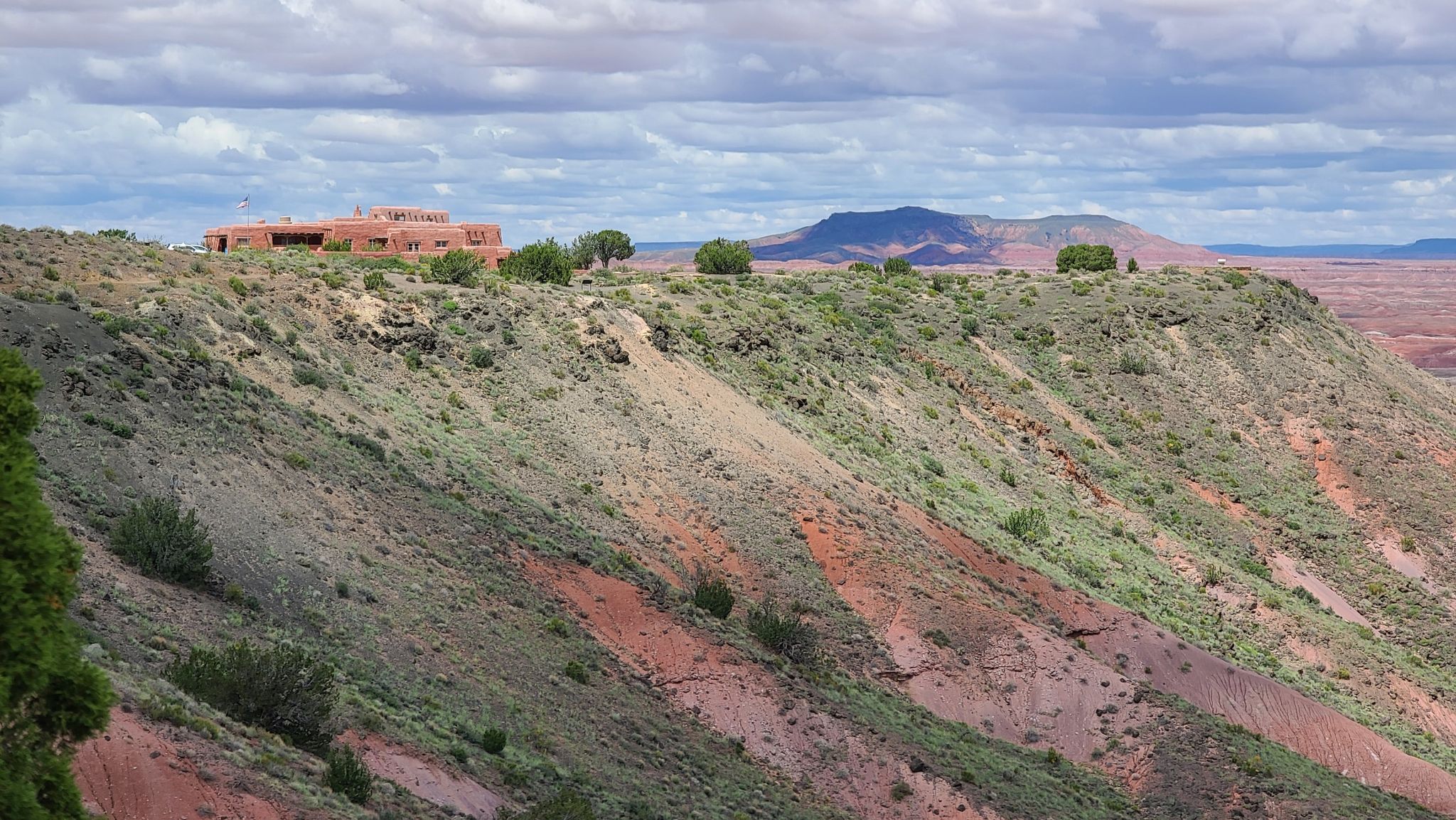 Distant red adobe building on a green and red hillside under a cloudy sky. - Painted Desert Rim Trail