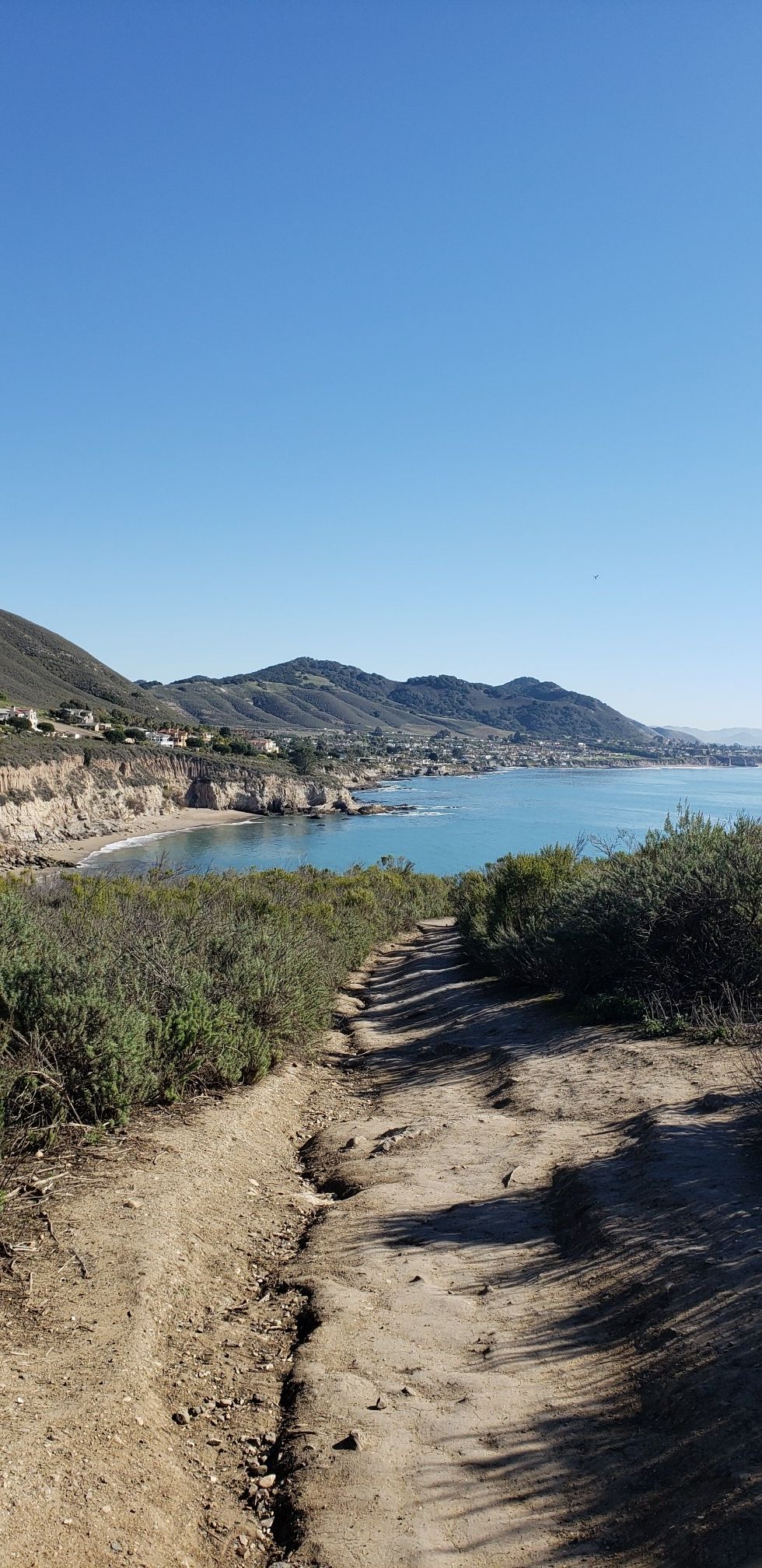 Sunny coastal landscape with a dirt path leading towards the ocean and mountains. - Shell Beach Bluff Trail
