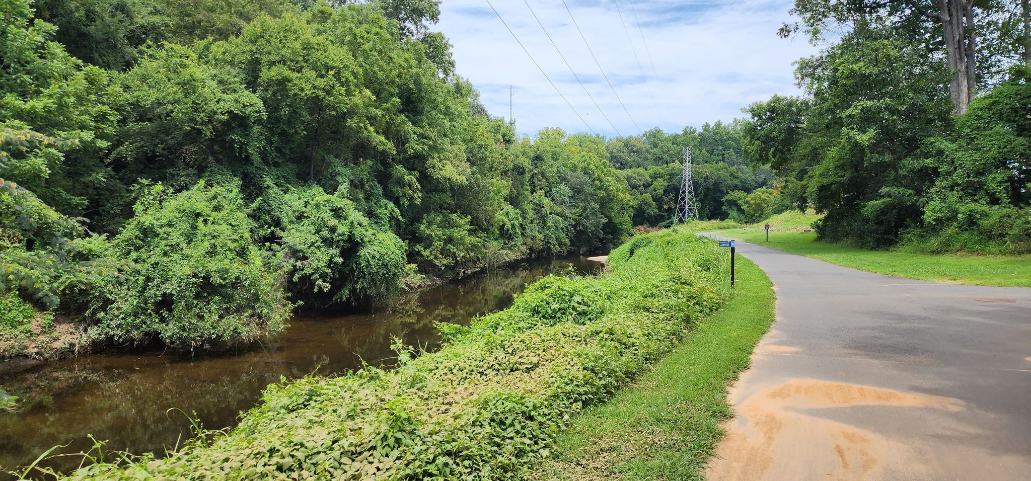 A narrow river flowing beside a paved path, surrounded by green trees. - Little Sugar Creek Greenway – Little Sugar Creek Greenway loop from Tyvola