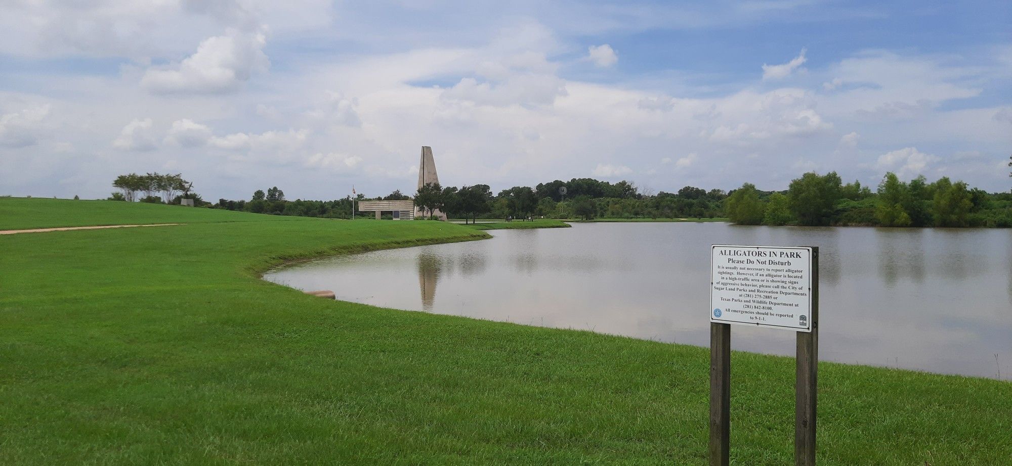 A calm lake with green grass, a distant monument, and a cloudy sky. - View of pond – Veterans Memorial Eagle loop from Trailhead