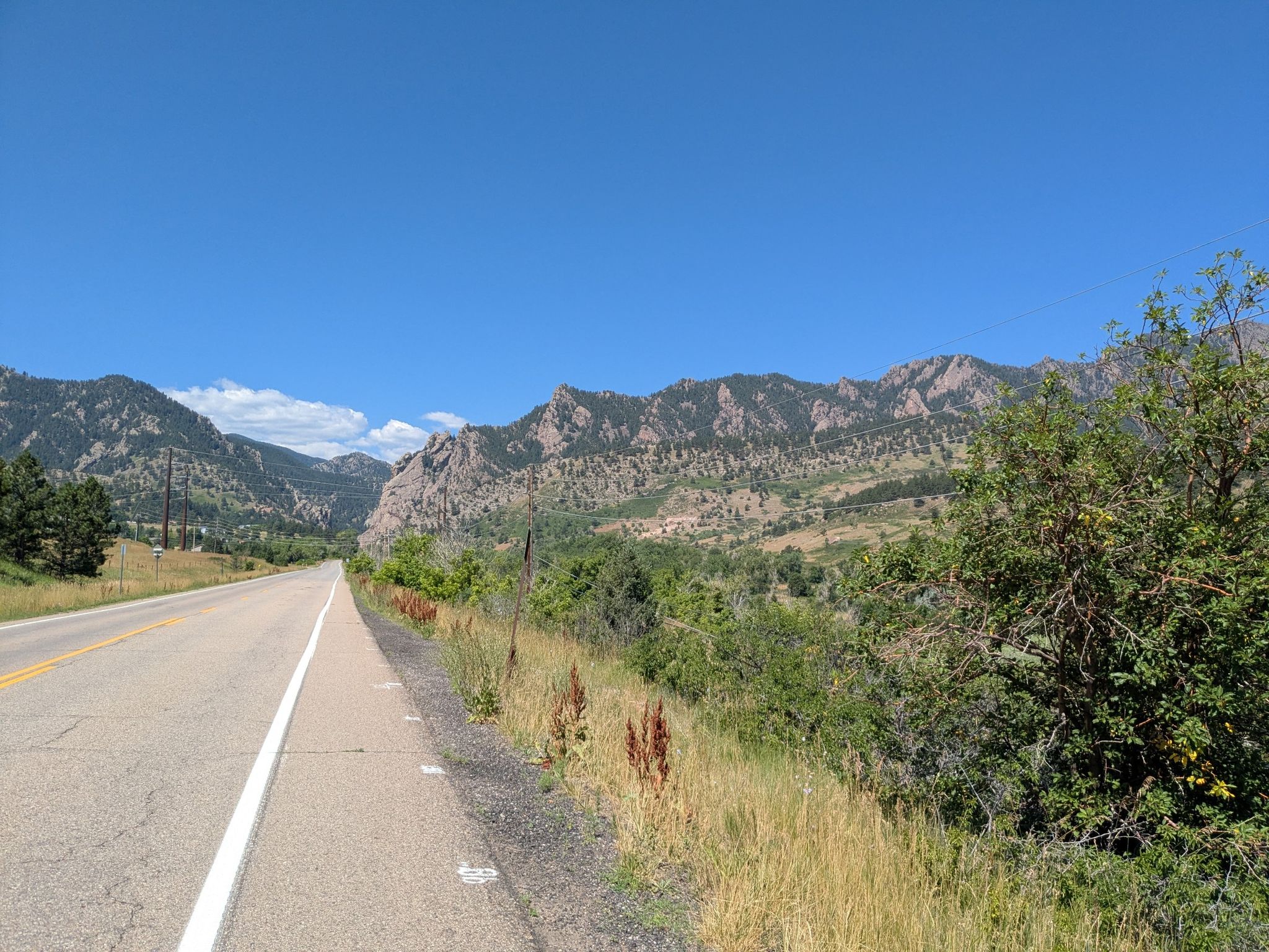 Paved road with a shoulder, green brush, and distant mountains under blue sky. - Shanahan Hill – Viele Lake loop from Culinary School of the Rockies