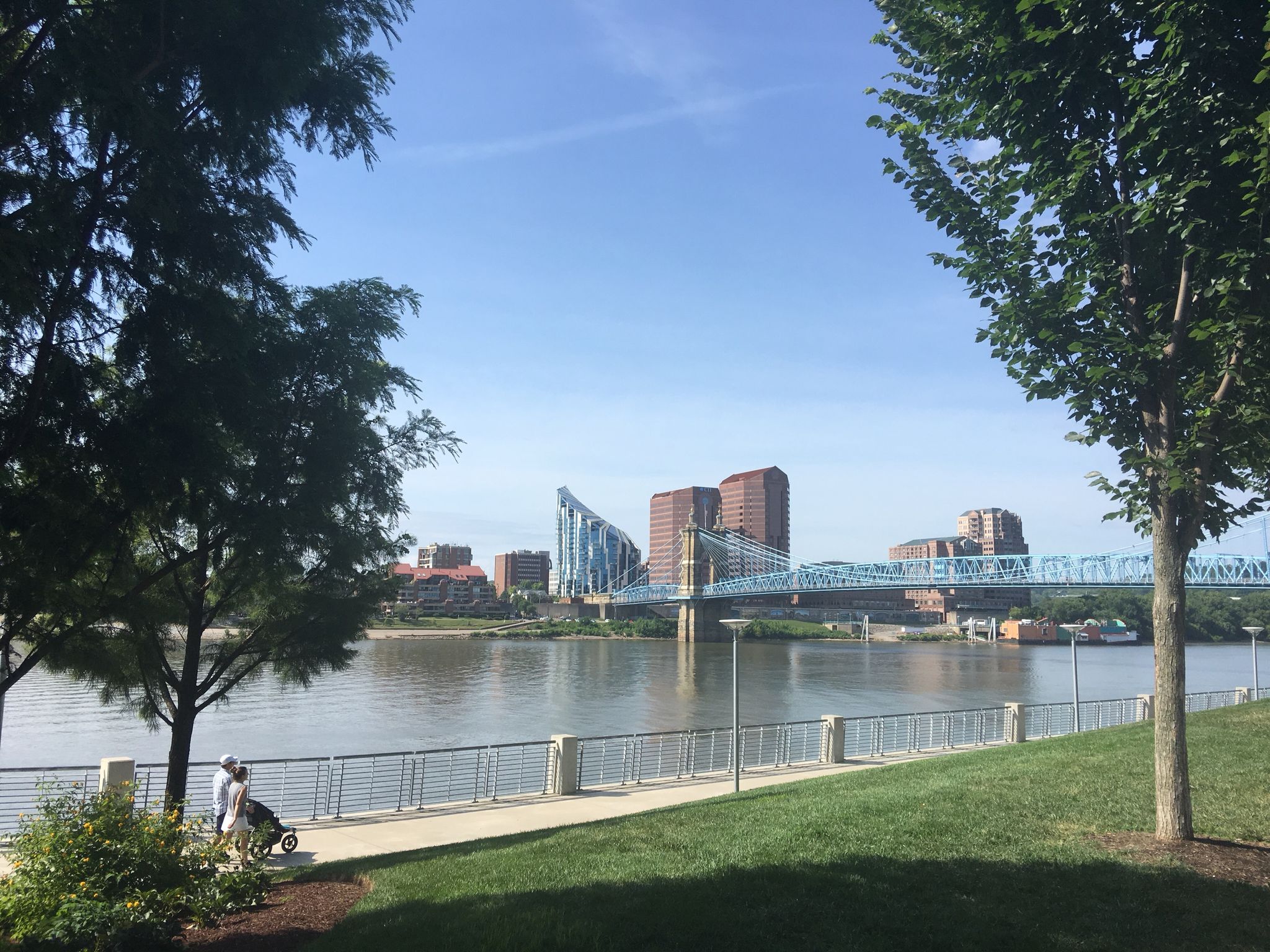 Sunny urban riverfront with a bridge, city buildings, and a person with a stroller. - Ohio to Erie Trail