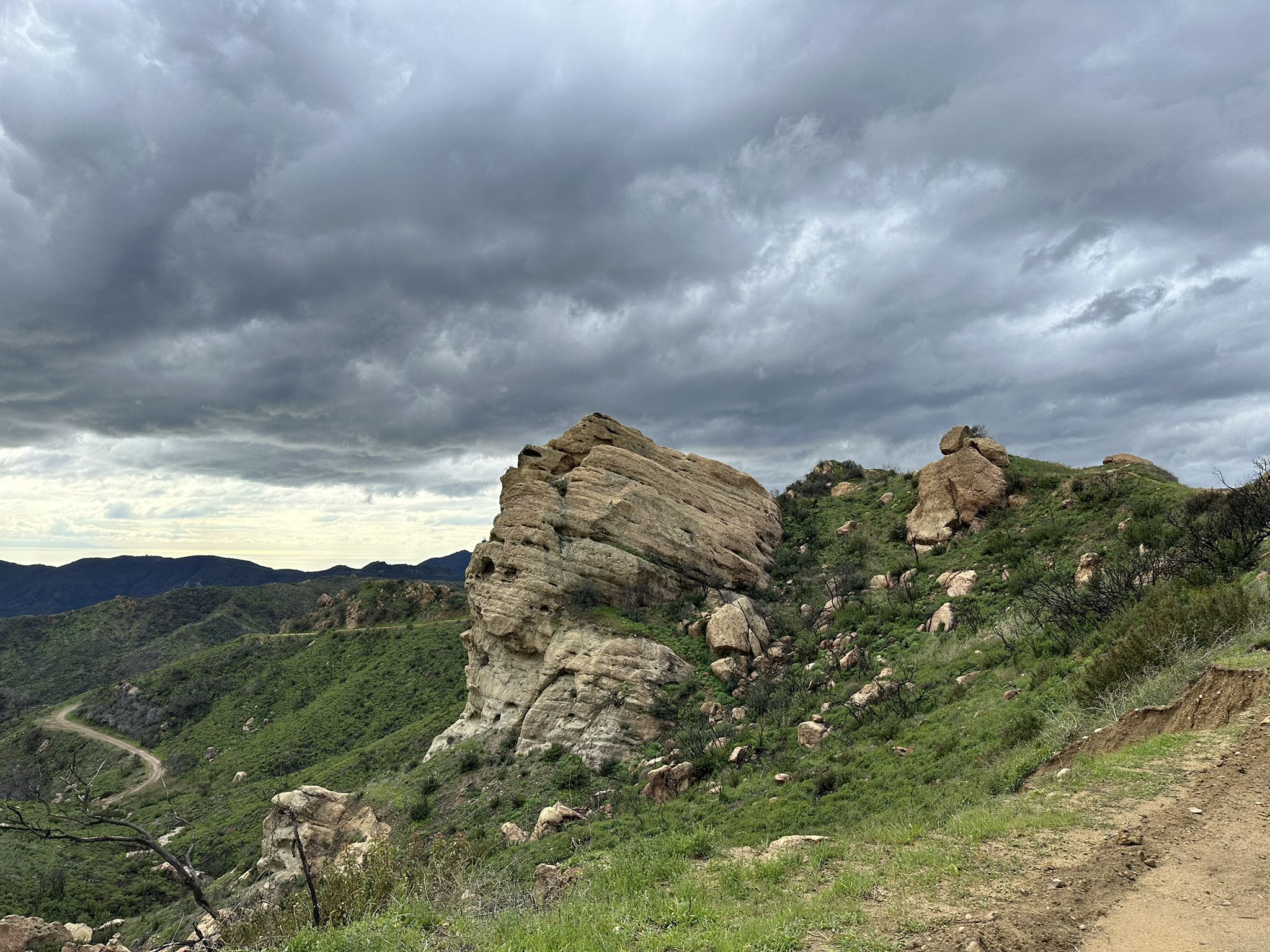 Large rock formation on green hillside under cloudy sky, winding road below. - East Topanga Fire Road Loop