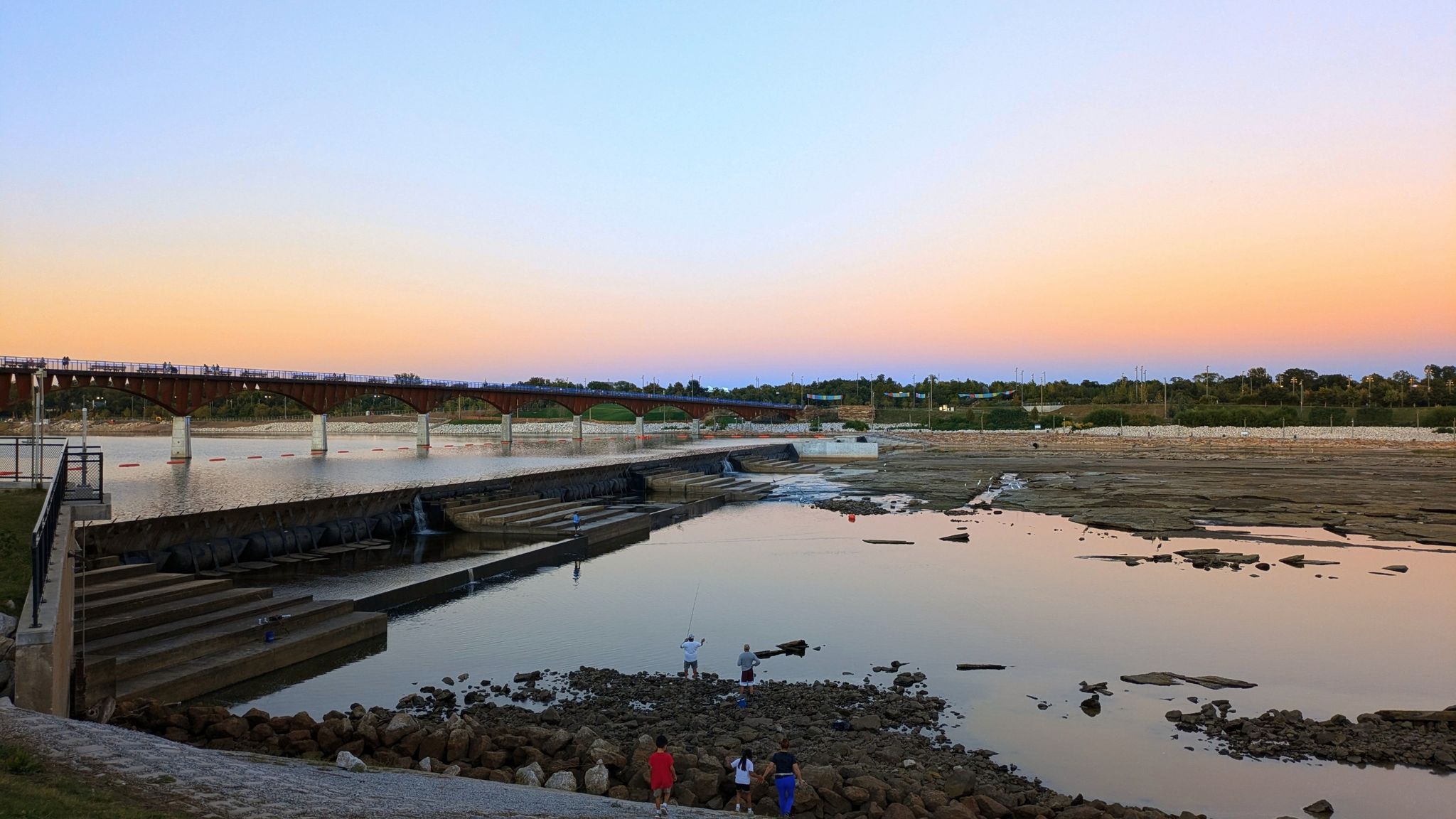 People fishing on a rocky riverbank at sunset, with a bridge in the distance. - The Gathering Place – Gathering Place Park loop from BancFirst