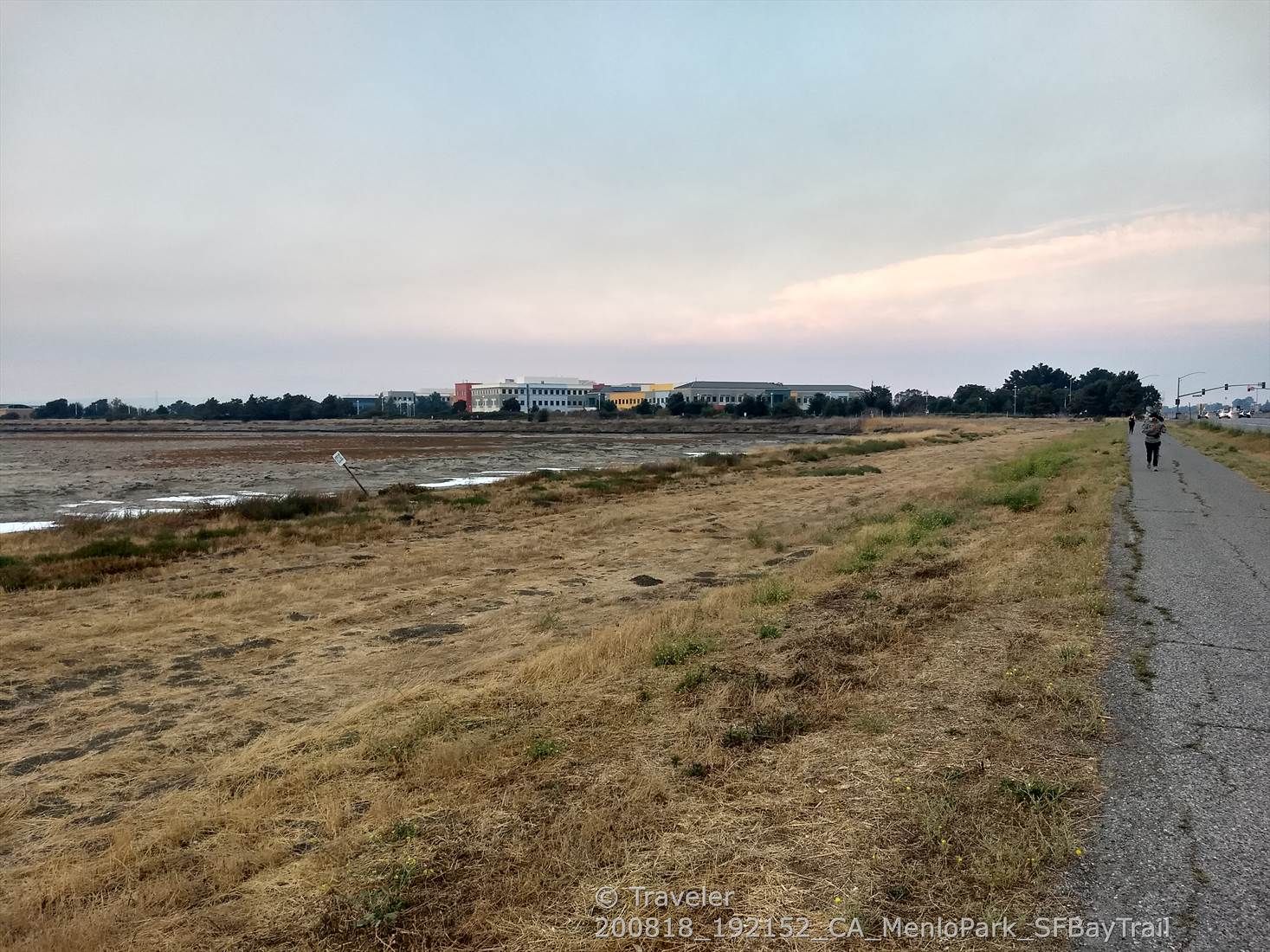 People walk on a paved path next to dry marshland and distant buildings. - San Francisco Bay Trail