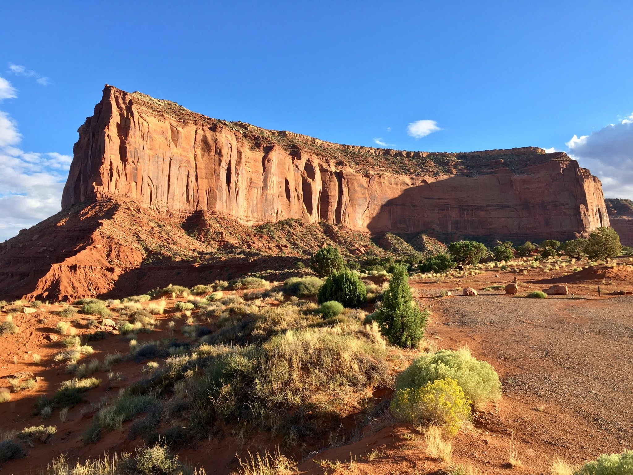 Massive red rock mesa with shadows, green desert plants, and a dirt road. - Monument Valley Campground Lee Cly Trail