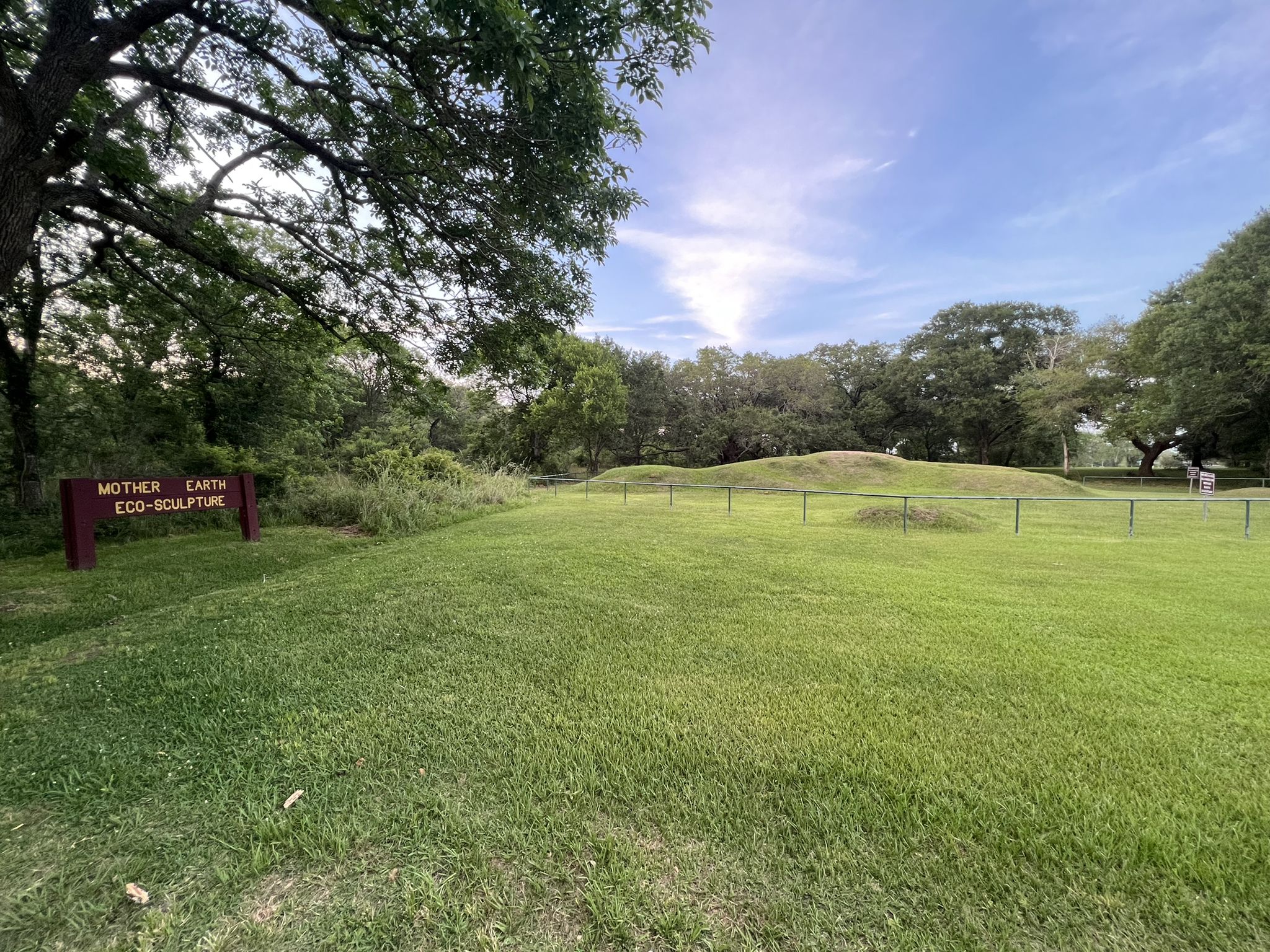 Sunny green field with a sign and distant mounds surrounded by trees. - Challenger Seven Park Loop
