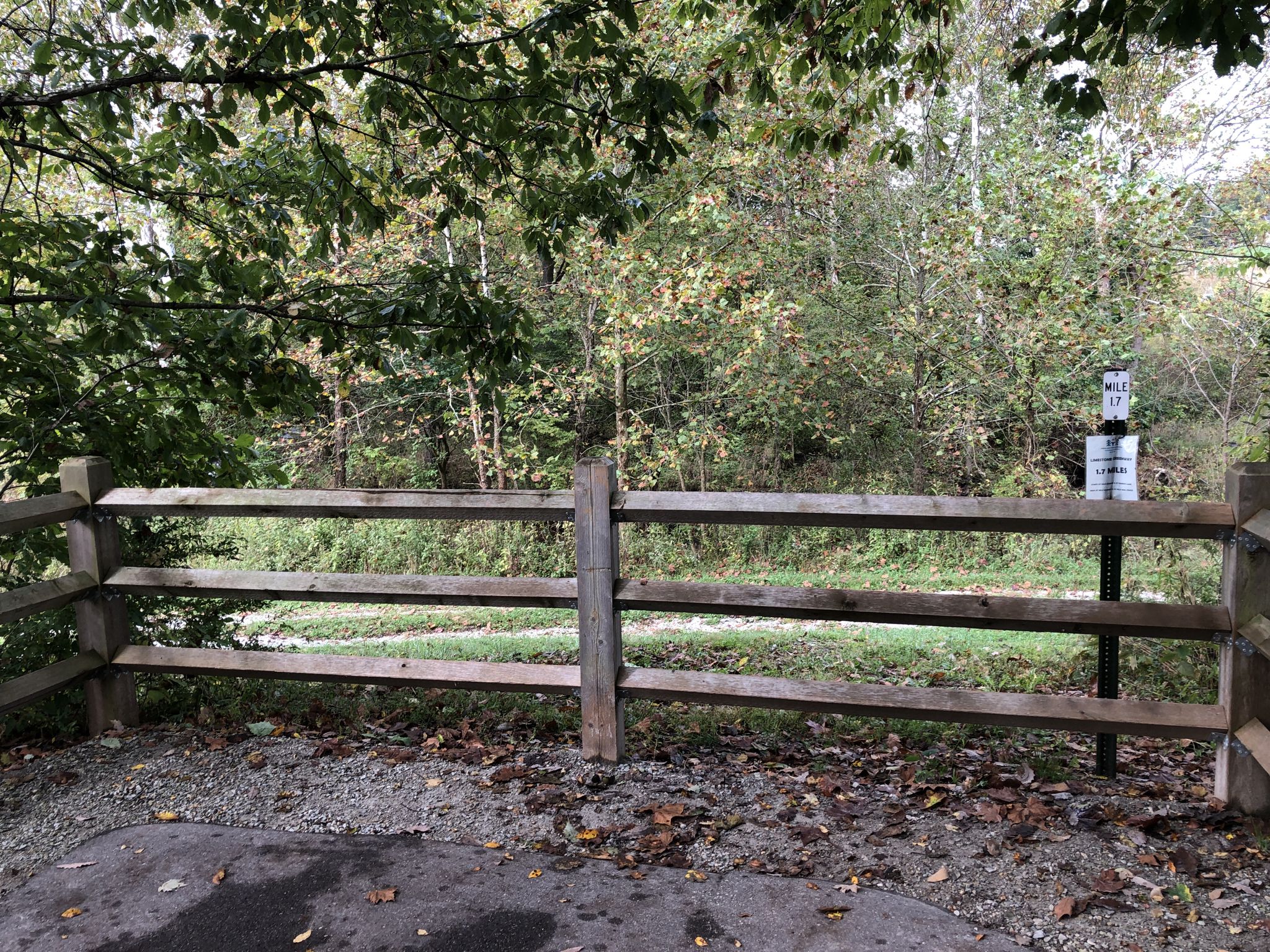 Wooden fence and sign next to a gravel path, surrounded by green trees. - Limestone Greenway