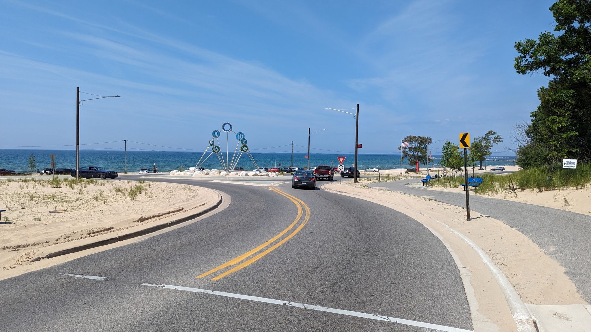 A curving road leads to a sunny beach with blue water and parked cars. - Lakeshore Trail, Lake Michigan – Muskegon Lake loop from Montague