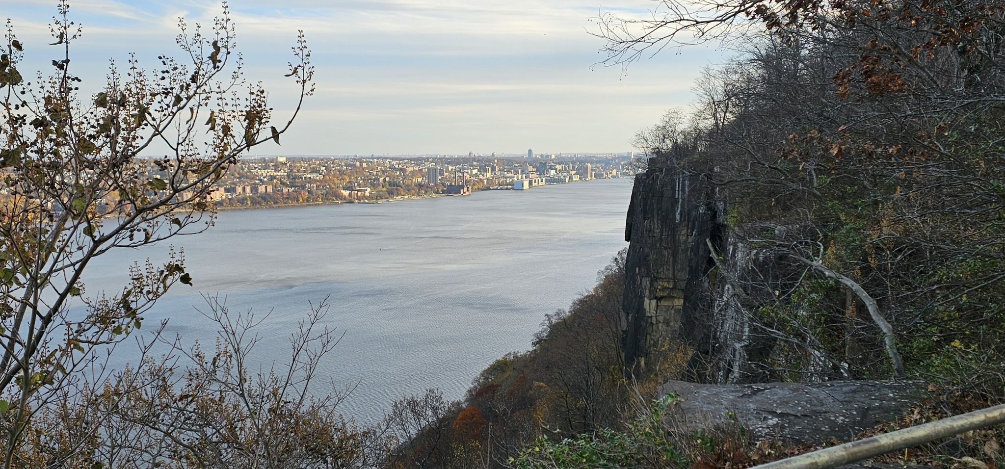 Overcast view of a wide river, distant city, and rocky cliff with bare trees. - Long Path and Shore Trail Loop