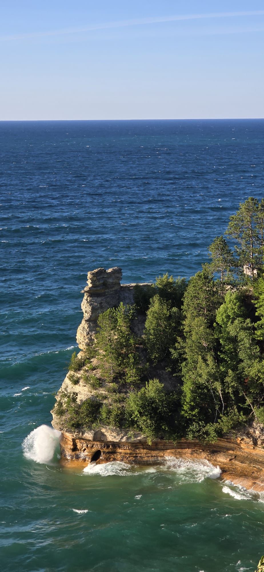 Sunny view of a rocky cliff with trees, waves crashing into the base. - Miners Castle – Miners Beach loop from Cliffs Camp