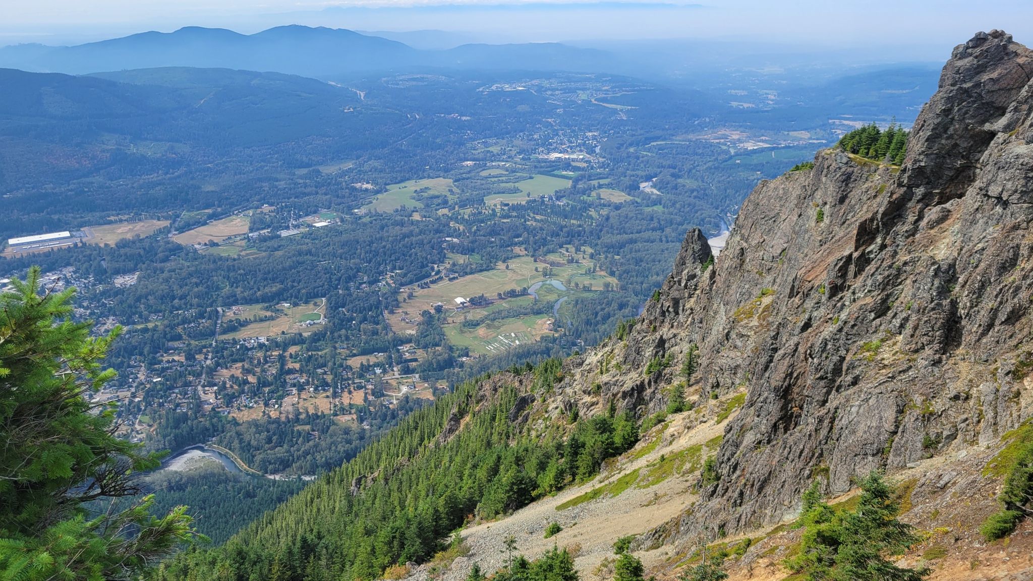 Steep rocky mountain slope overlooks a vast forested valley and distant town. - Mount Si Viewpoint – Little Si Trail Viewpoint loop from Riverpoint