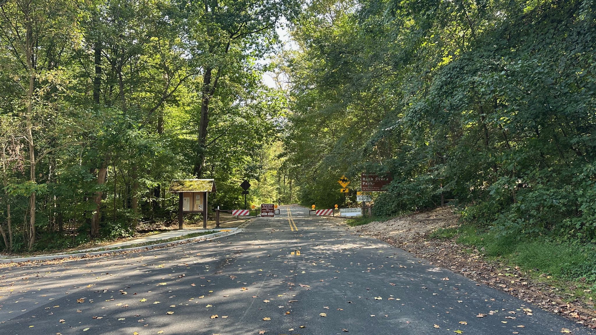 A paved road is blocked by barriers, surrounded by dense forest. - Meadowbrook Stables loop from Forest Glen