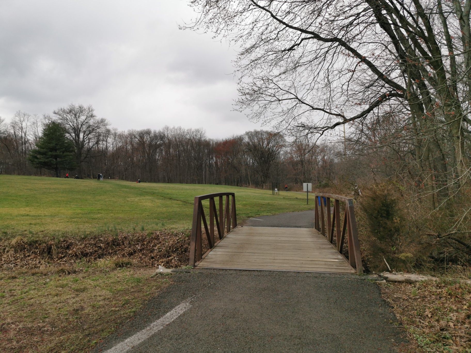 A wooden bridge crosses a paved path next to a grassy field under an overcast sky. - Oak Ridge Park Trail