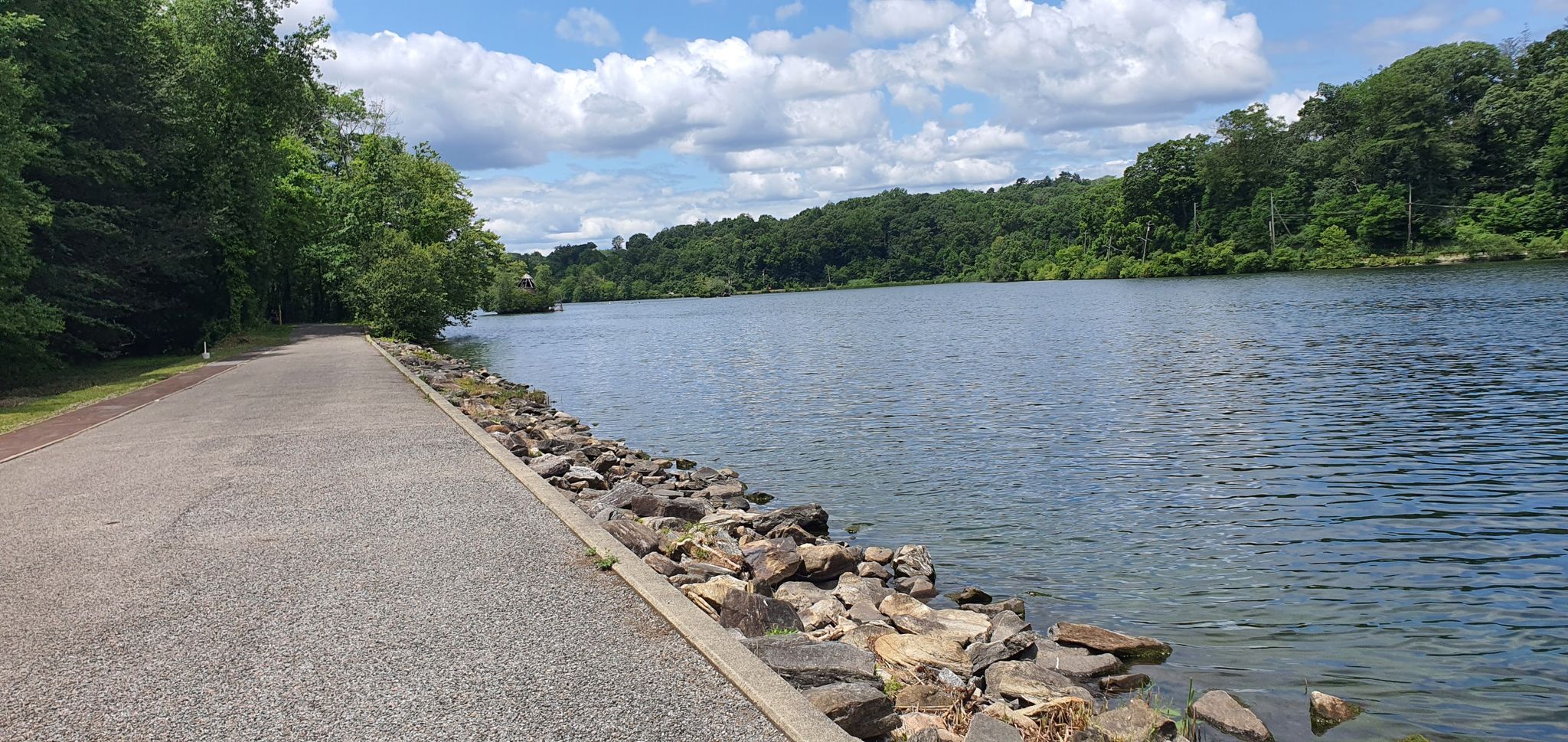 Paved path alongside a wide lake with rocky shore and distant green hills. - Oradell Reservoir – Tarrytown Reservoir loop from Oradell