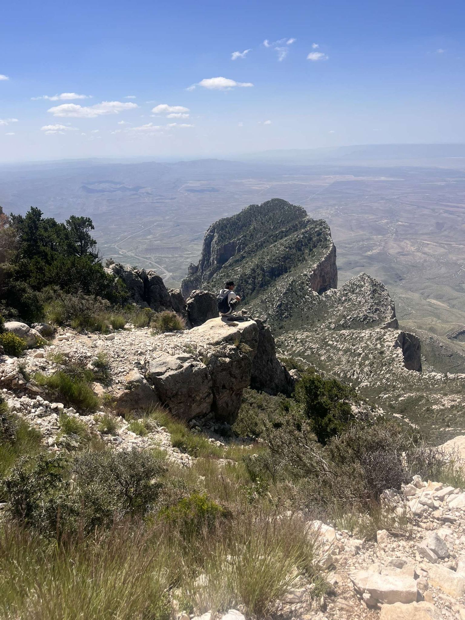 Person sits on a rocky outcrop, overlooking a vast mountain and valley landscape. - Pine Springs Visitor Center – Ruins of Pinery Station loop from Pine Springs
