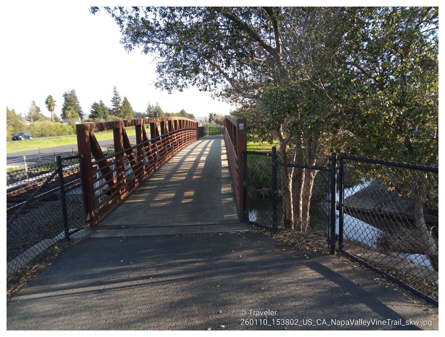 A metal pedestrian bridge with shadows, next to a fence and trees, on a sunny day. - Napa Valley Vine Trail – Andretti Winery loop from Union