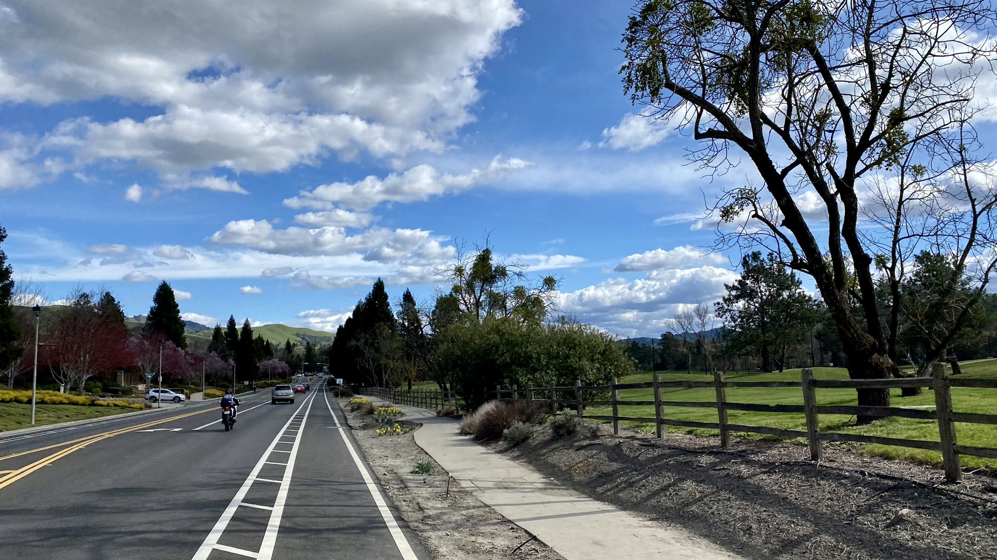 A road with a bike lane, cars, and a motorcycle, next to a green field. - Moraga Commons Park – Moraga Ranch & Café loop from Moraga