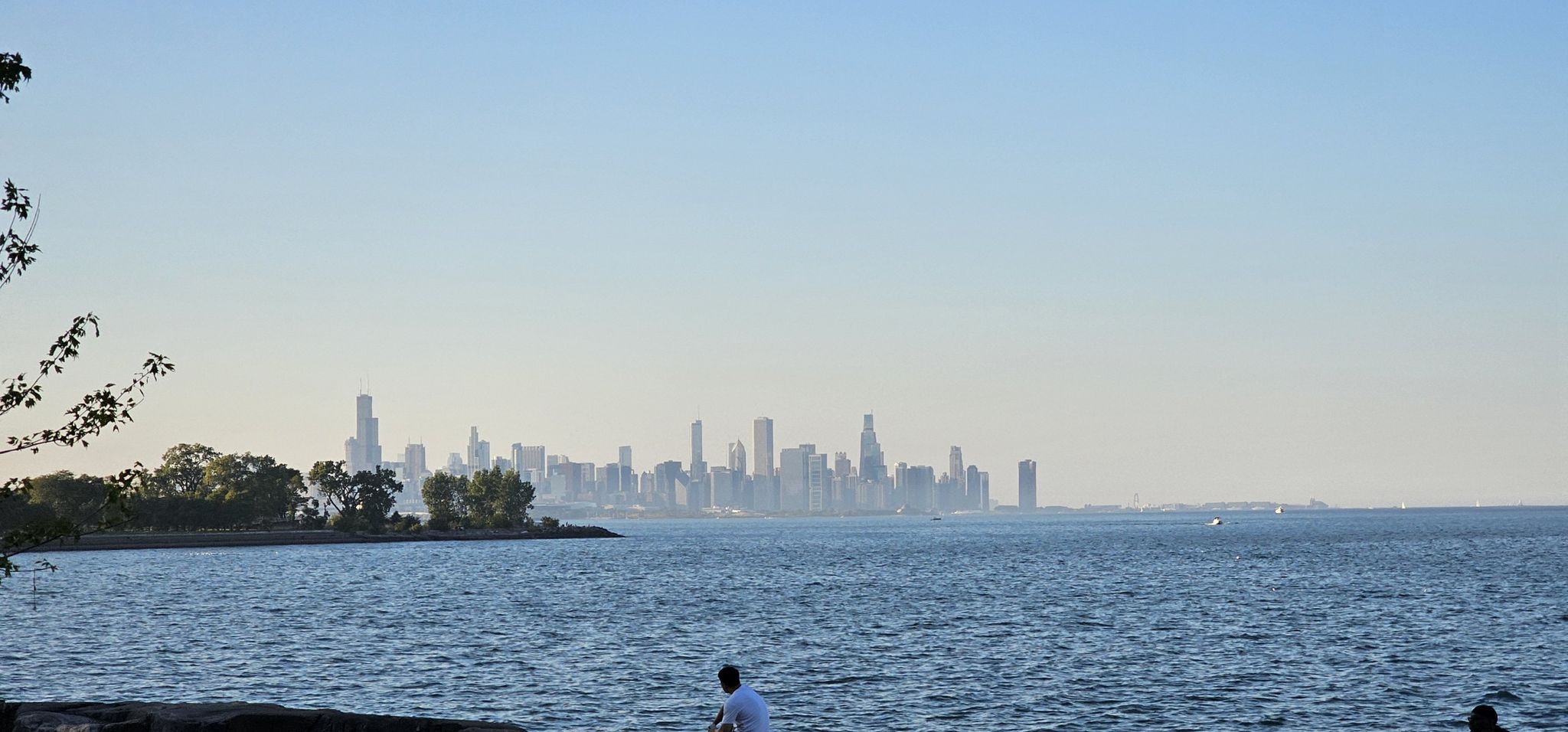 Person sits by water, viewing a distant city skyline under clear skies. - Hyde Park – Model Yacht Basin loop from The University of Chicago