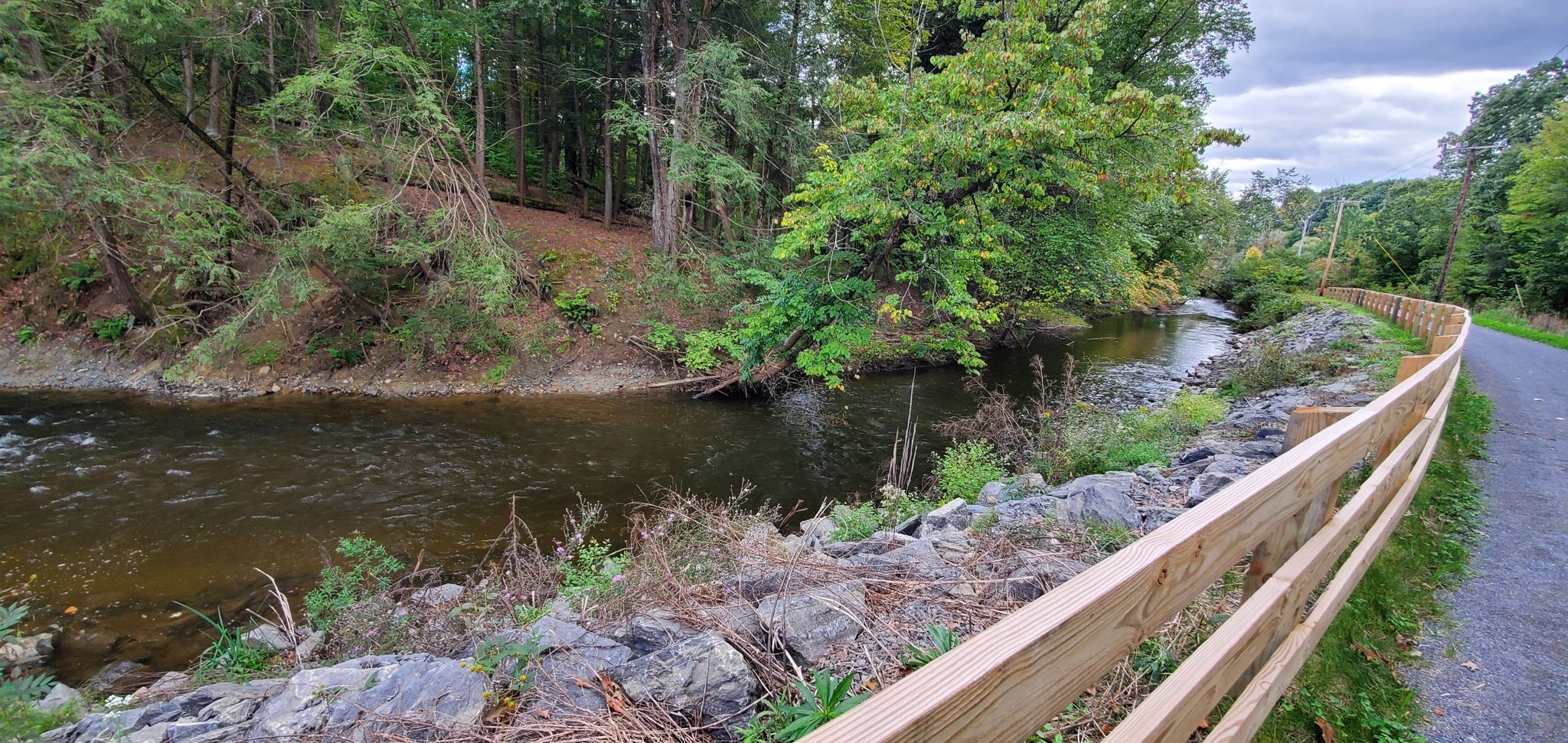 River flowing alongside a paved path and wooden fence in a forest. - Bike loop from Valatie