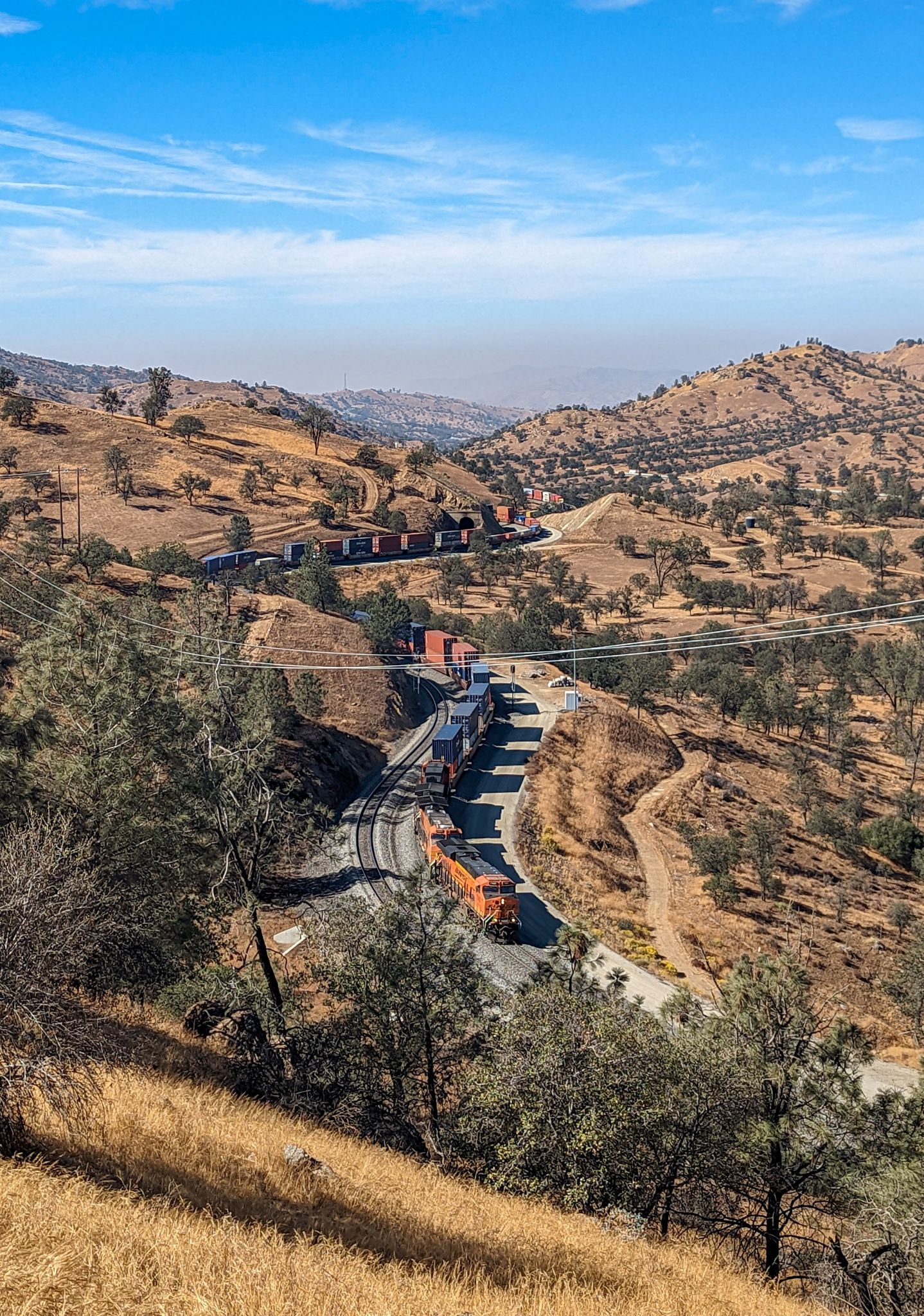 Train winds through dry, hilly terrain and scattered trees on a sunny day. - Tehachapi Railroad loop