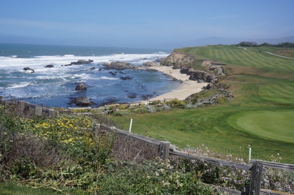 Sunny view of a rocky beach, ocean, and green golf course with a wooden fence. - Miramontes Point – Wavecrest Open Space Preserve loop from Johnston Ranch