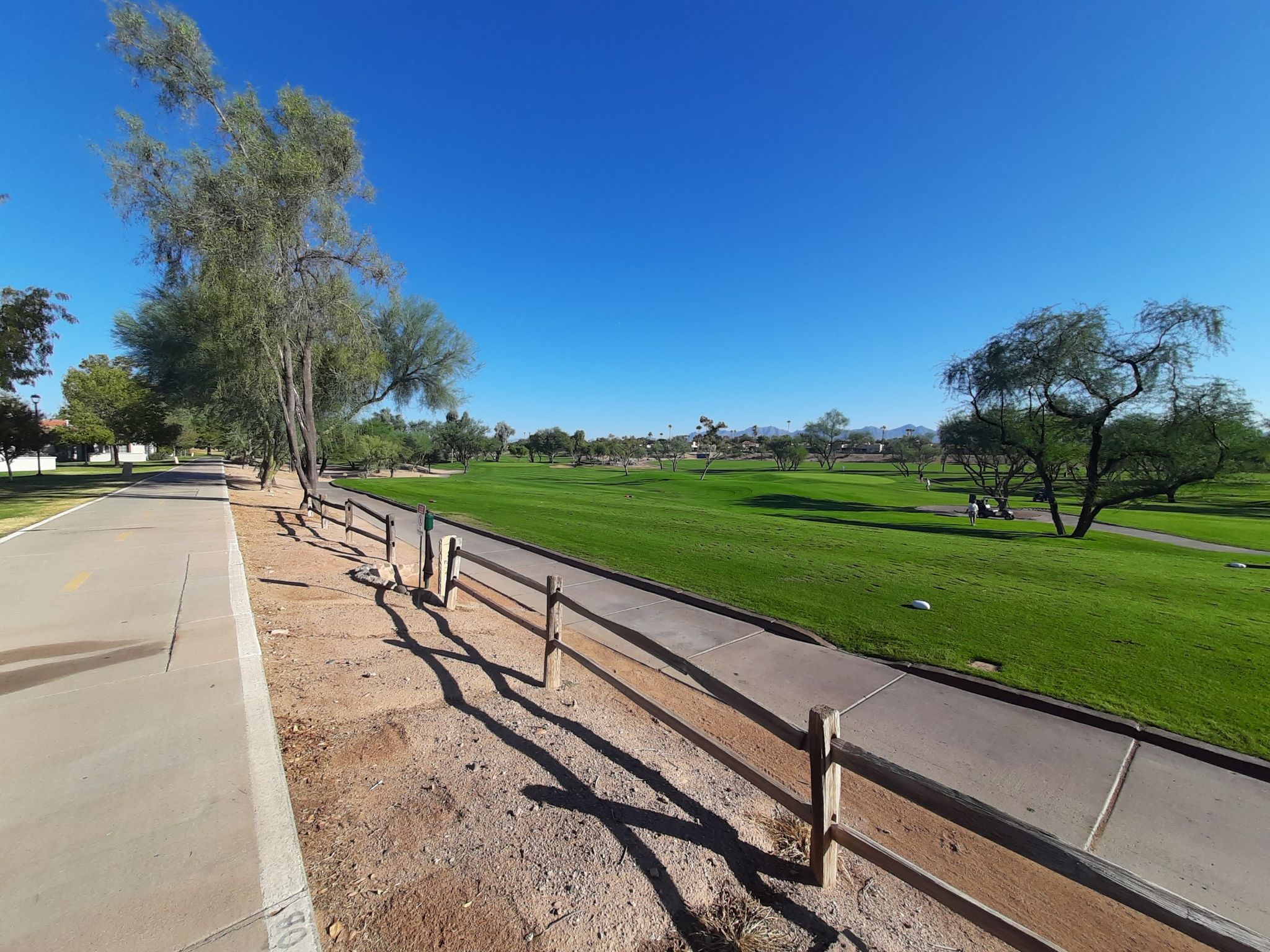 A wooden fence borders a paved path next to a green lawn. - Scenic view of Camelback – Sun Circle Trail Canal Path loop from McCormick Stillman Railroad Park
