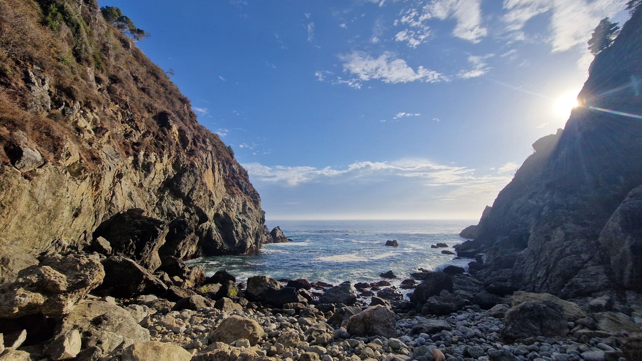 Sun shines brightly over a rocky ocean cove between two steep cliffs. - Partington Cove Trail