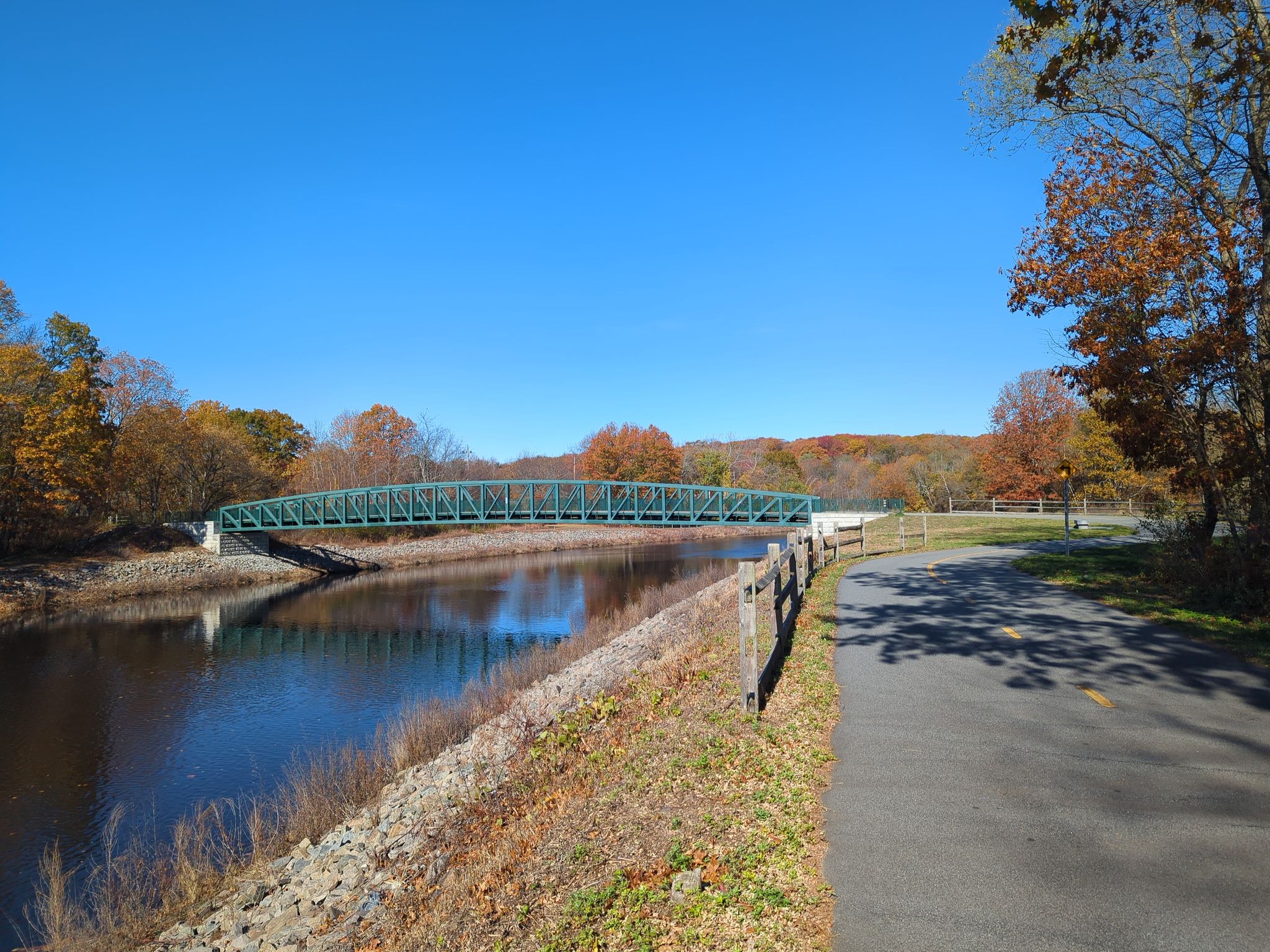 A green bridge spans a river with autumn trees under a bright blue sky. - Blackstone River Bikeway