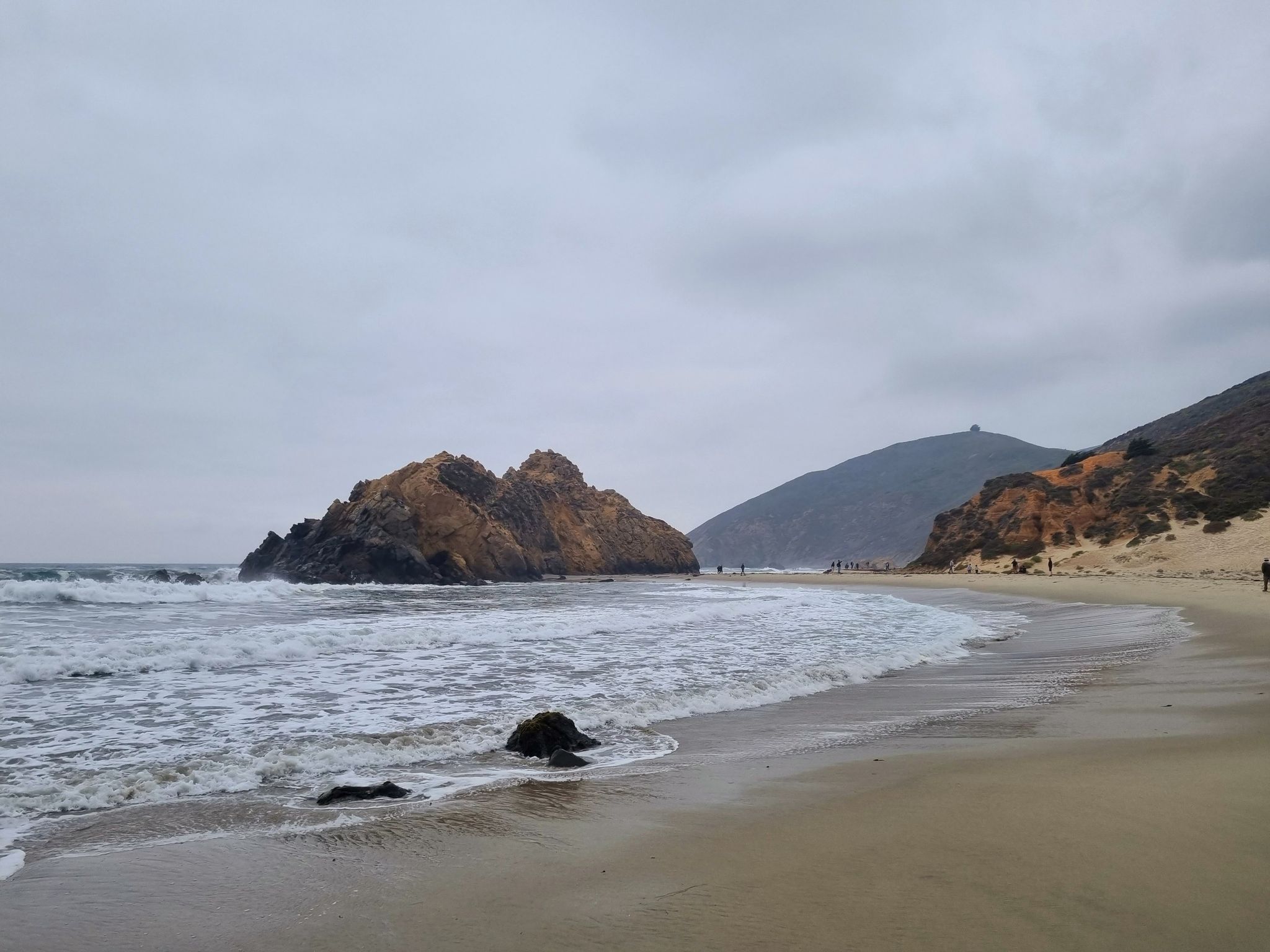 Overcast beach scene with waves, a large rock, and distant hills. - Pfeiffer Beach