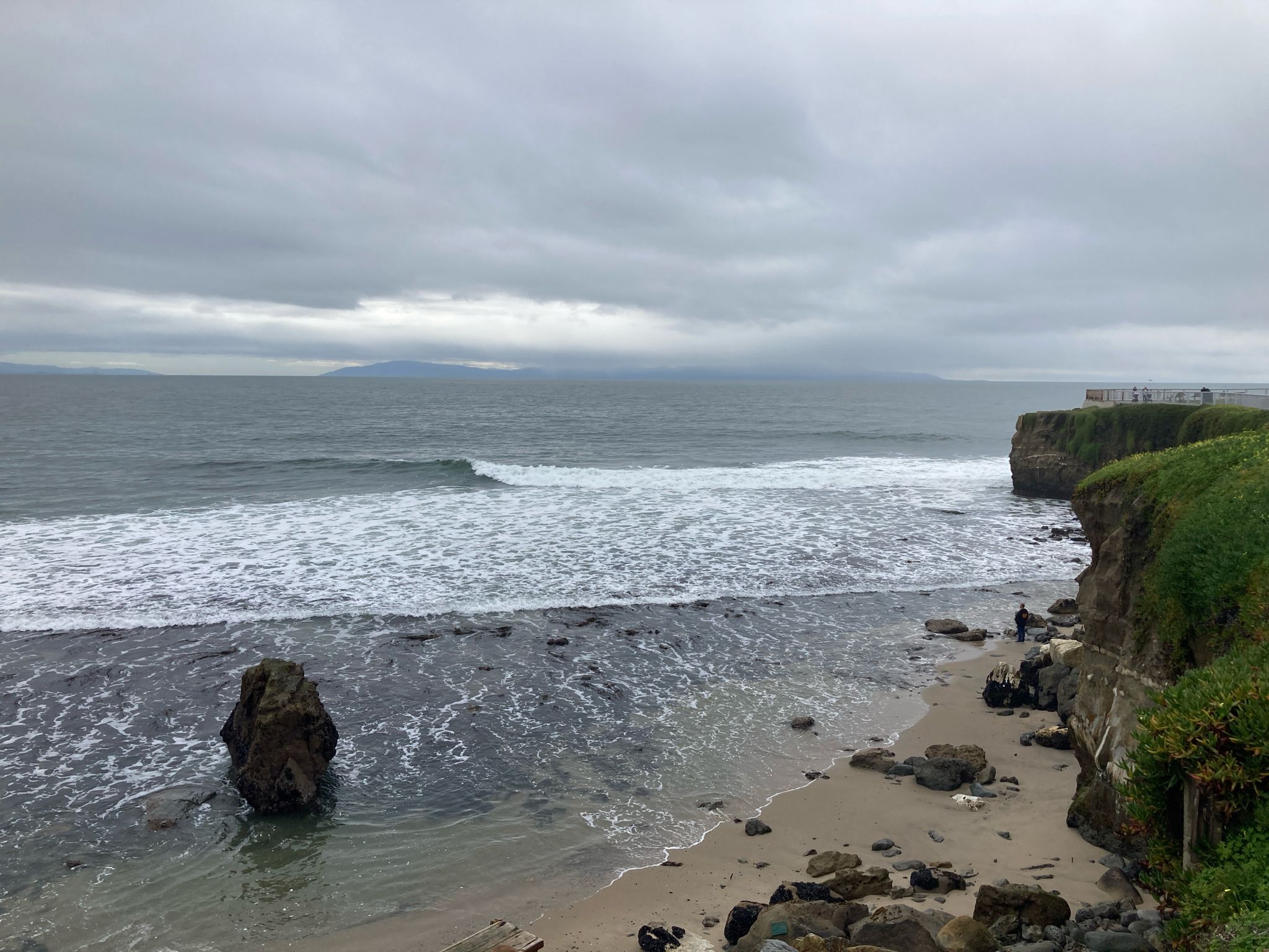 Overcast beach scene with breaking waves, a large rock, and distant cliffs. - Surfer Statue – Santa Cruz Lighthouse loop from Santa Cruz