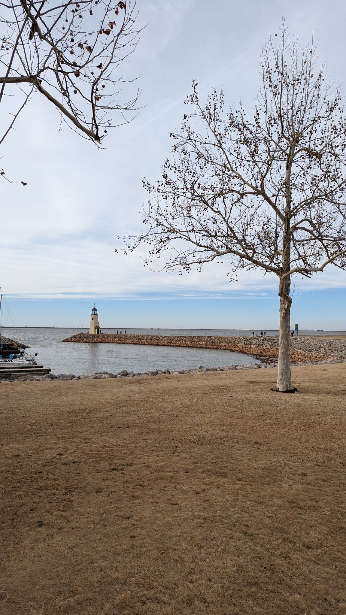 A distant lighthouse on a rocky breakwater, with bare trees and brown grass. - Lake Hefner Lighthouse – Bert Cooper Trails loop from Hefner Junior High School