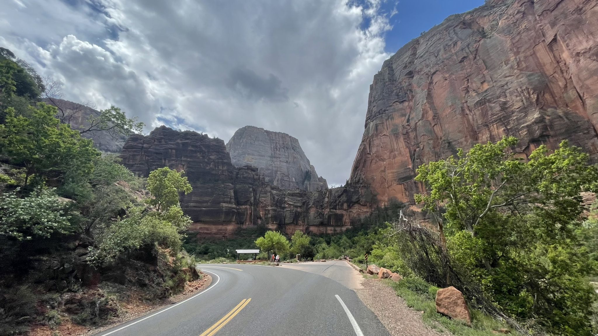 Winding road through a canyon with towering rock formations under cloudy skies. - Zion National Park Scenic Drive