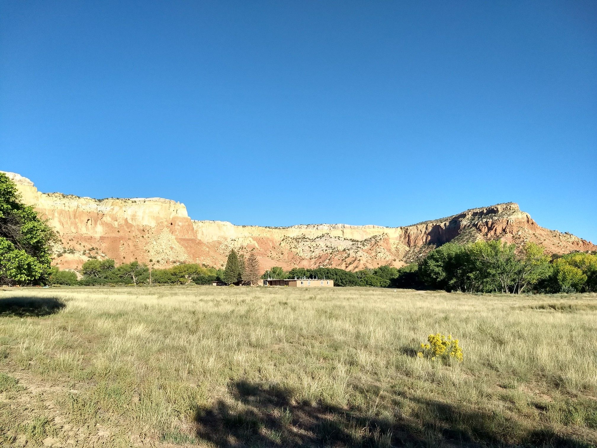 Wide grassy field with distant buildings and large rock cliffs under a clear sky. - Kitchen Mesa Trail