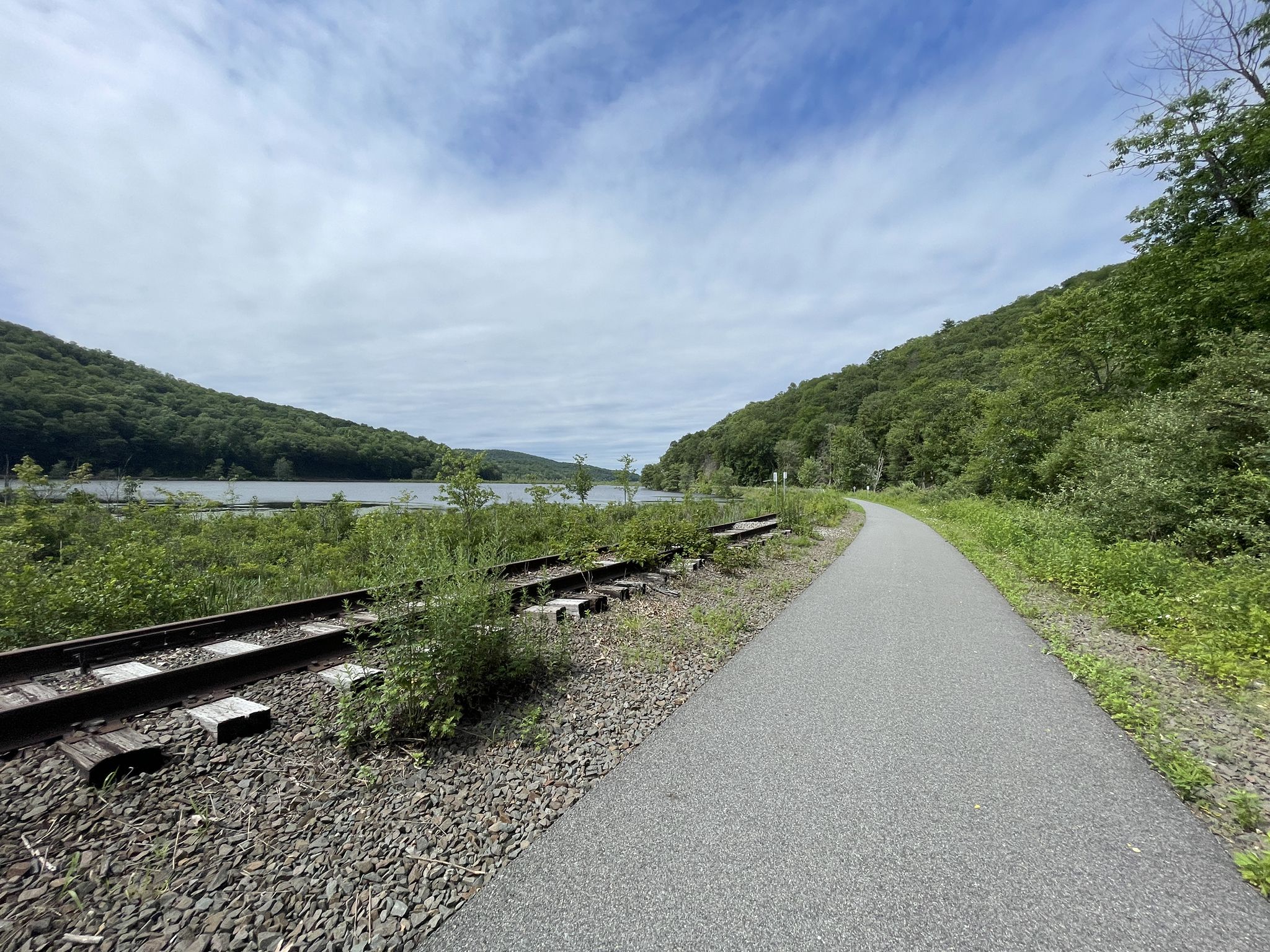 A paved path and train tracks run alongside a lake, surrounded by green hills. - Maybrook Trailway loop from Brewster Hill