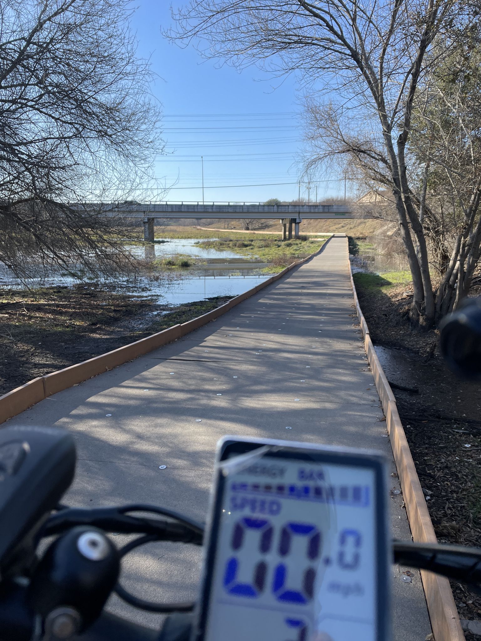 A bike's dashboard view of a paved path over water, with a bridge in the distance. - Morningstar Boardwalk – Morningstar Boardwalk loop from MacArthur High School